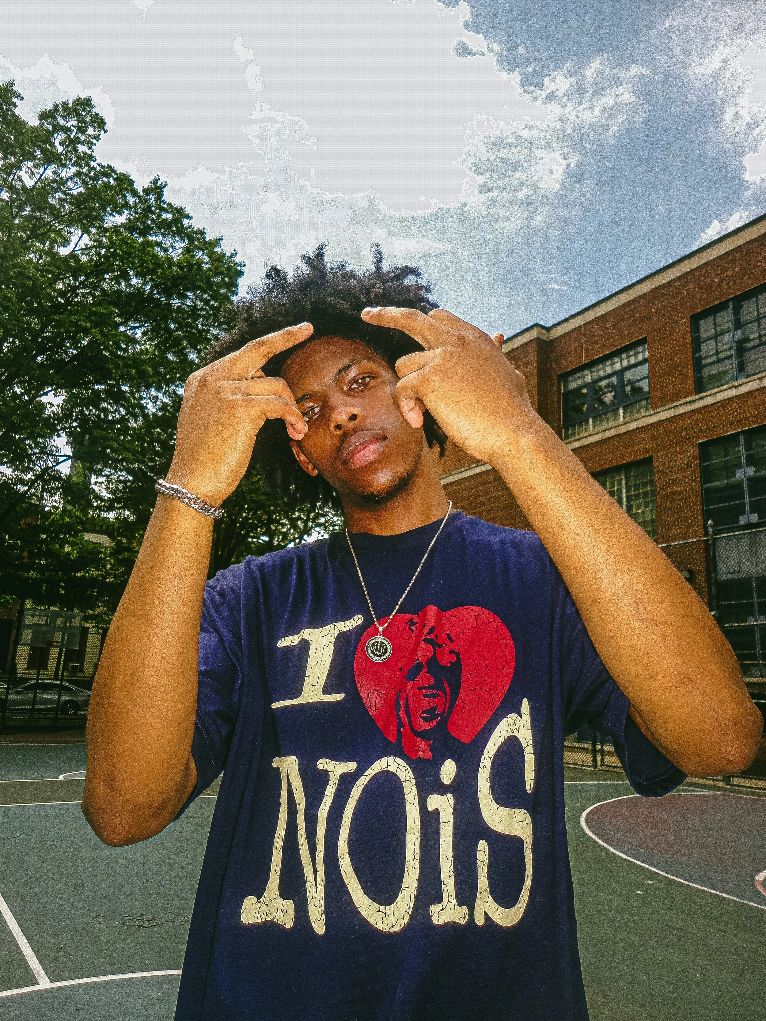 Young man with curly hair making hand gestures on a basketball court outdoors, wearing a dark T-shirt with a graphic and text, a necklace, and a bracelet, with trees and a brick building in the background.