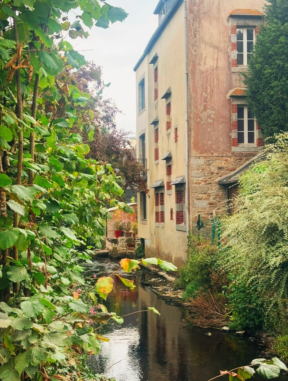 A hospitable town in Brittany, France with a river flowing past old stone and brick buildings set amongst lush, green foliage.