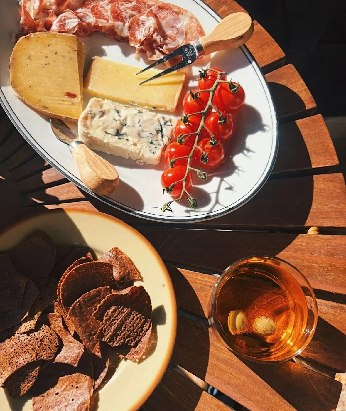 An al fresco apéro setting on a wood table in Brittany, France—fresh french cheese, sliced cured meats, a vine of cherry tomatoes, a plate of blé noir crisps, and a spritz with green olives by The Blackfjord for present gathering.