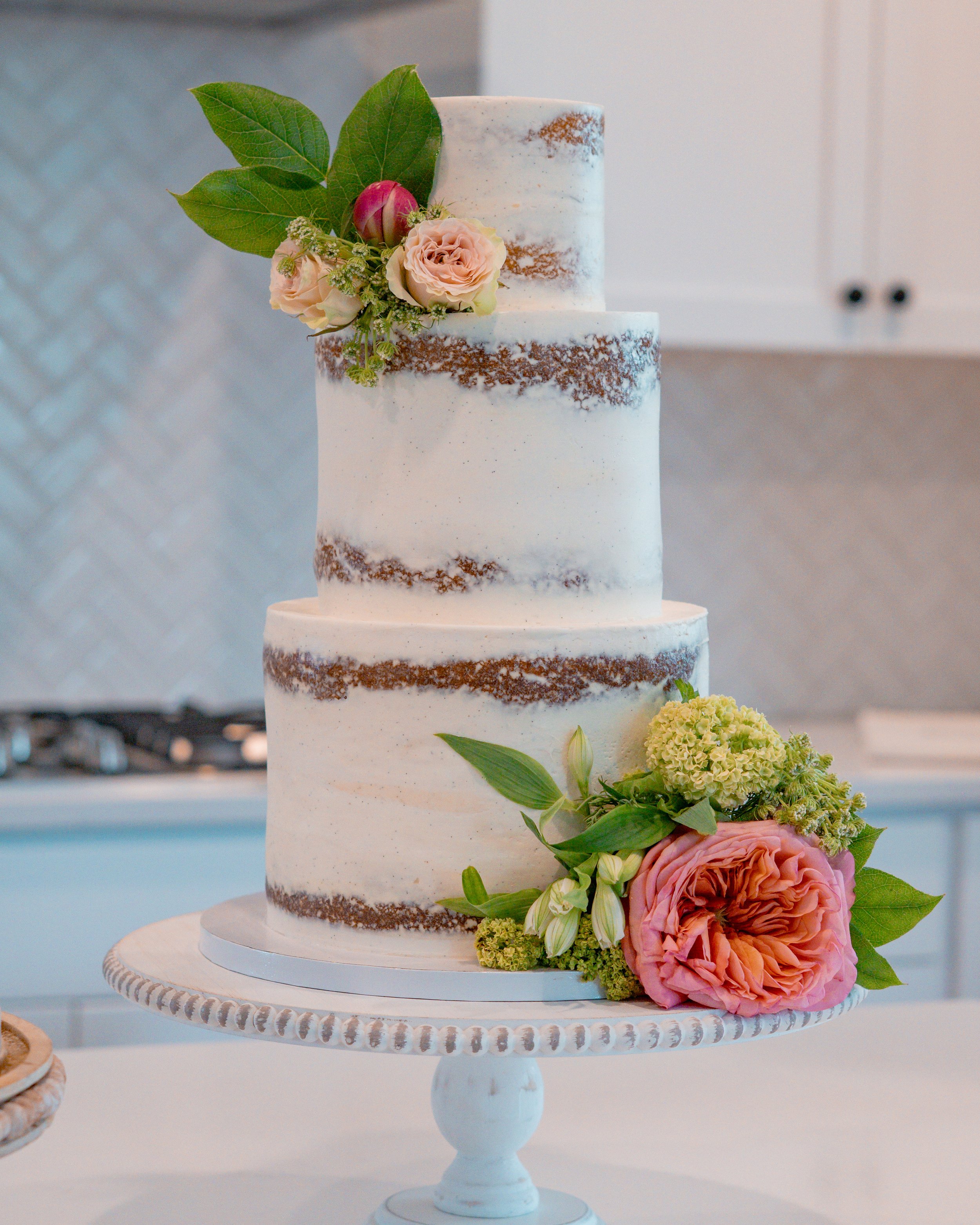 A tall, three-tiered white wedding cake with semi-naked icing and floral decorations, including pink roses, a large pink peony, white lilies, green leaves, and small white filler flowers, placed on a decorative white cake stand.