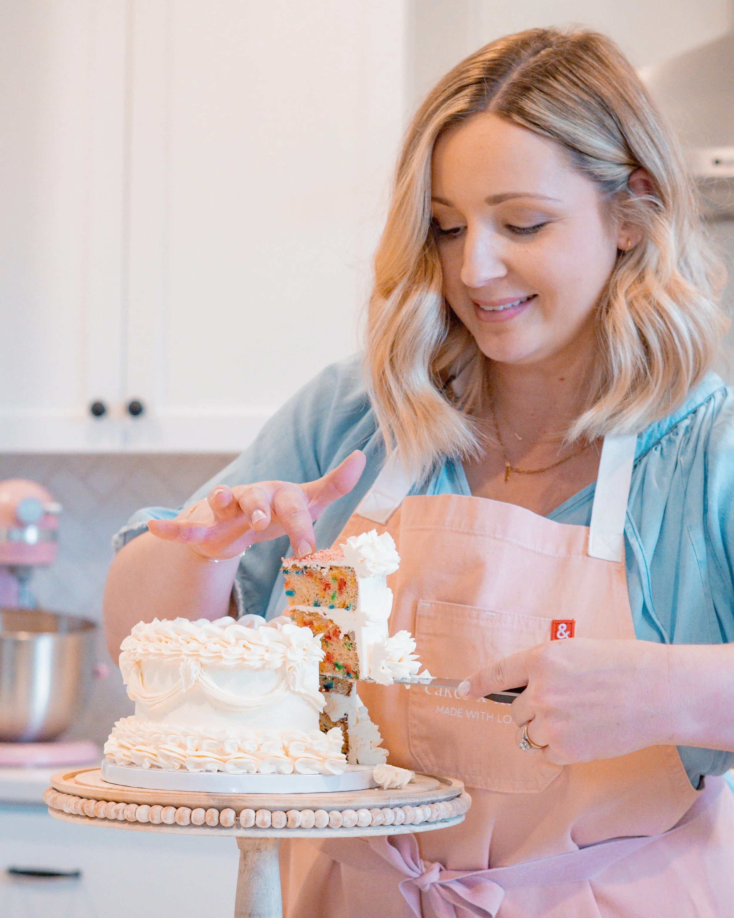 A woman wearing a pink apron is decorating a tiered birthday cake with white frosting and colorful sprinkles. She is smiling and looking down at the cake, which is on a wooden cake stand in a kitchen with white cabinets in the background.
