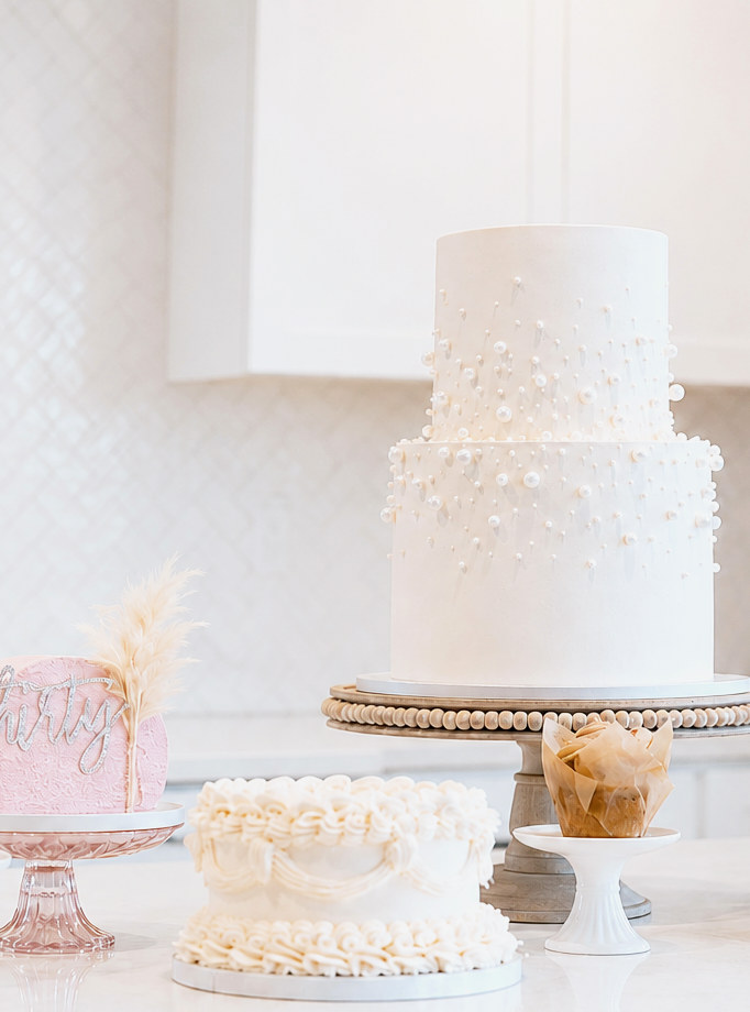 Three cakes on a table, including a large white tiered cake with pearl decorations, a pink cake with a feather and silver lettering, and a white cake with ornate piping, along with a cupcake in a wrappers.