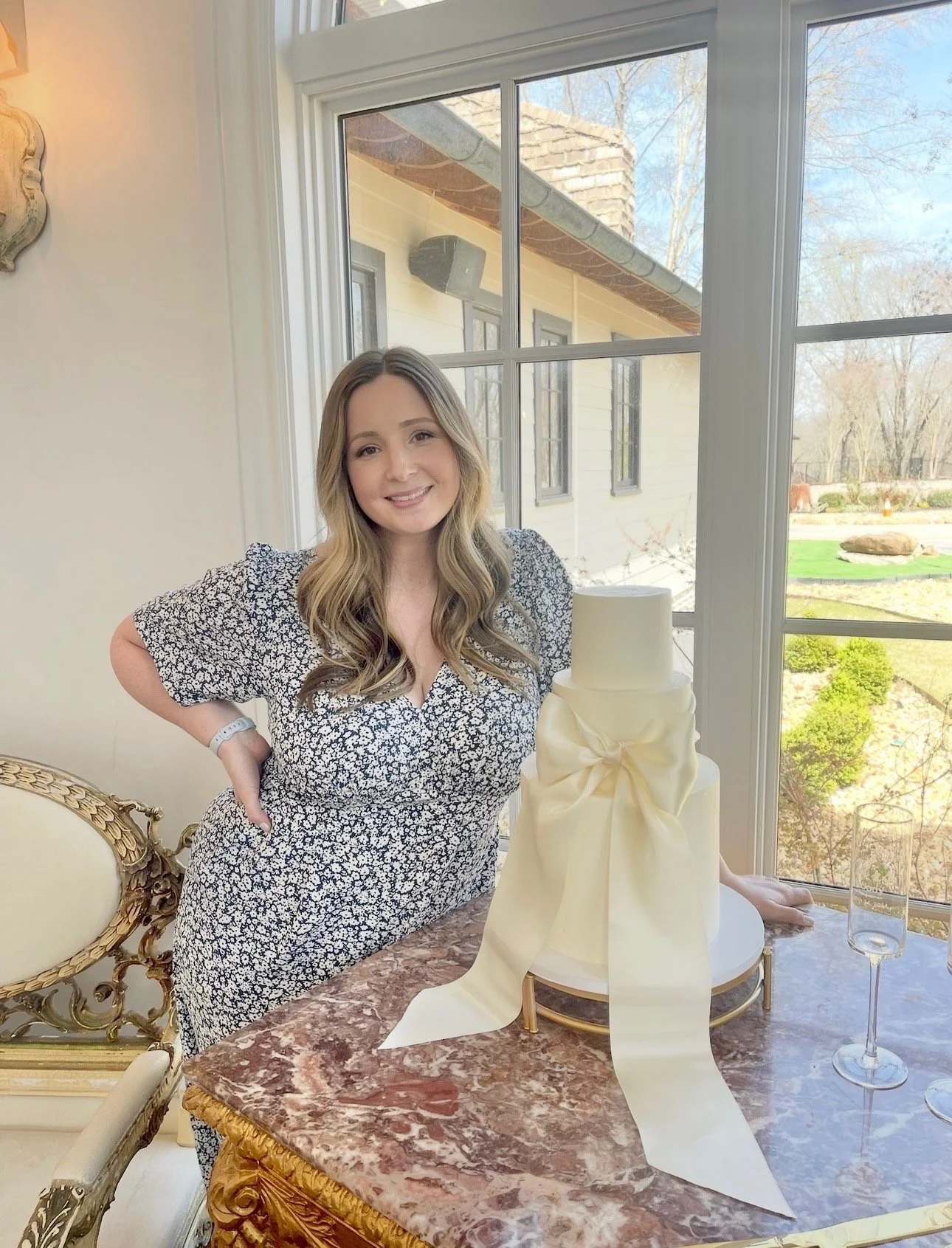 A woman with long wavy hair and a floral black and white dress is smiling and posing next to a white cake with a cream-colored ribbon on a marble table in front of a large window showing an outdoor scene with trees and a house.