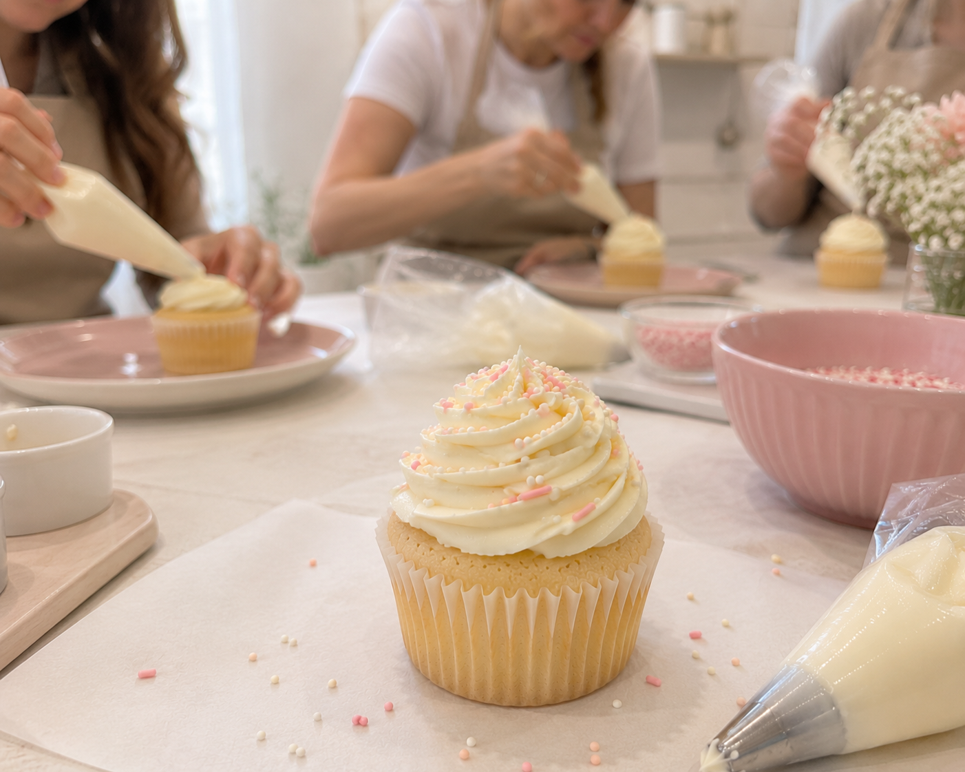 A cupcake with white frosting and pink and white sprinkles on a baking sheet. In the background, people are decorating cupcakes with frosting, surrounded by bowls of pink and white icing, a piping bag, and flowers.