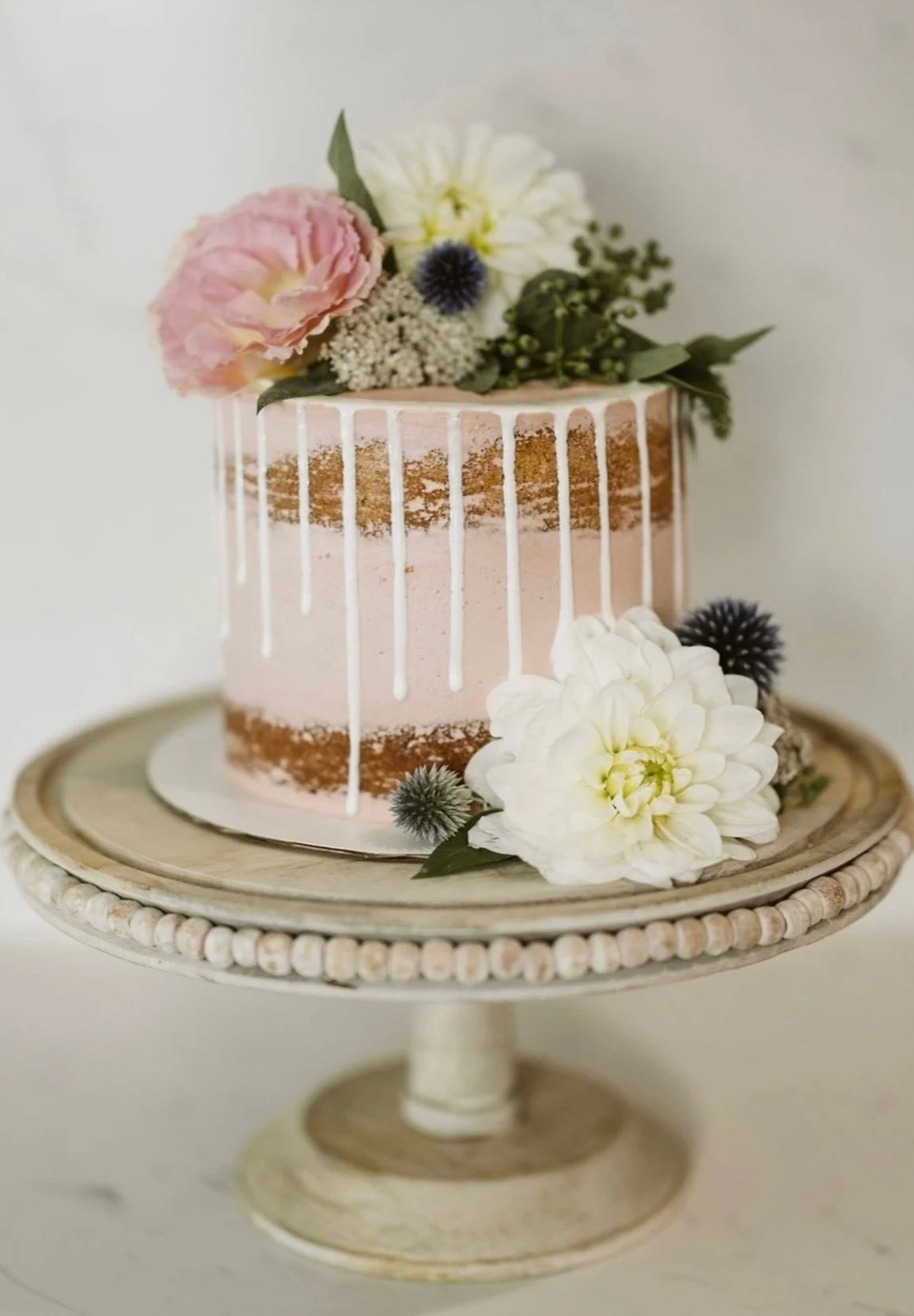 A pink semi-naked cake decorated with white drip icing, fresh flowers, and greenery on a rustic cake stand.