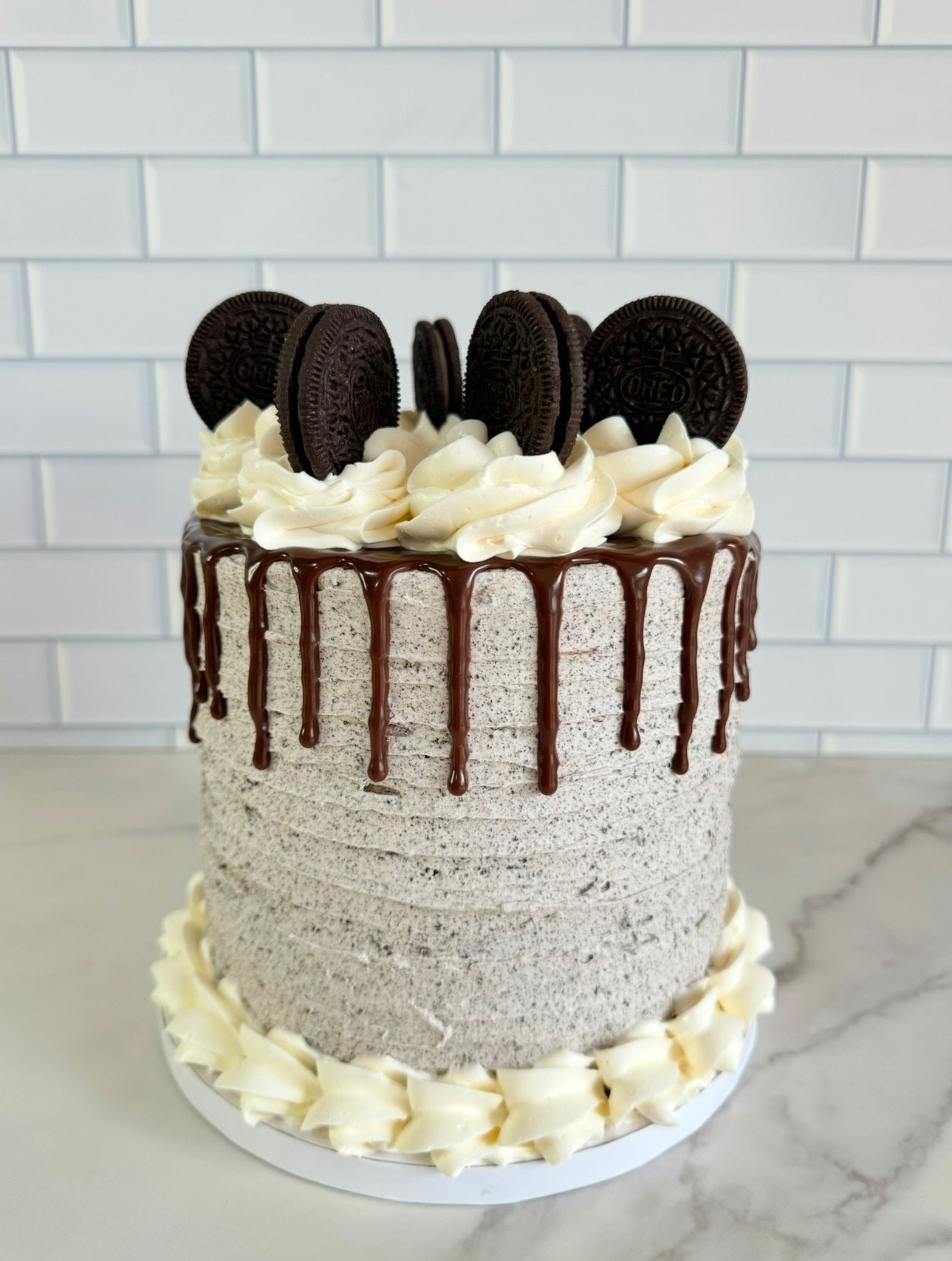 A tall birthday cake with a textured gray frosting, chocolate drip decoration, topped with Oreos and white whipped cream swirls, on a white cake board with white chocolate decoration around the base, against a white tiled background.