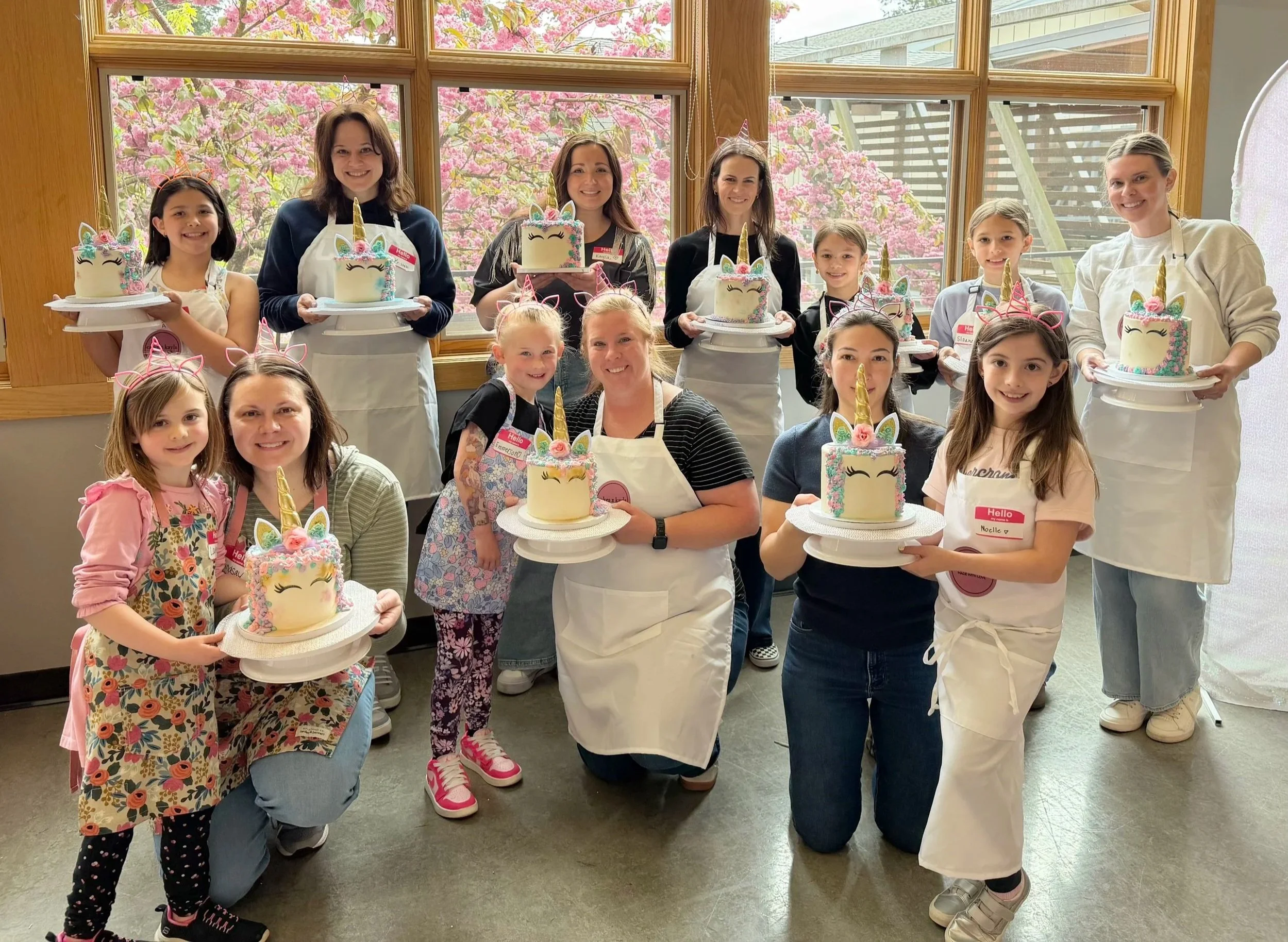 A group of children and adult women celebrating a unicorn-themed birthday party indoors with decorated cakes featuring unicorn faces and horn decorations, with pink blossoms visible outside the window.