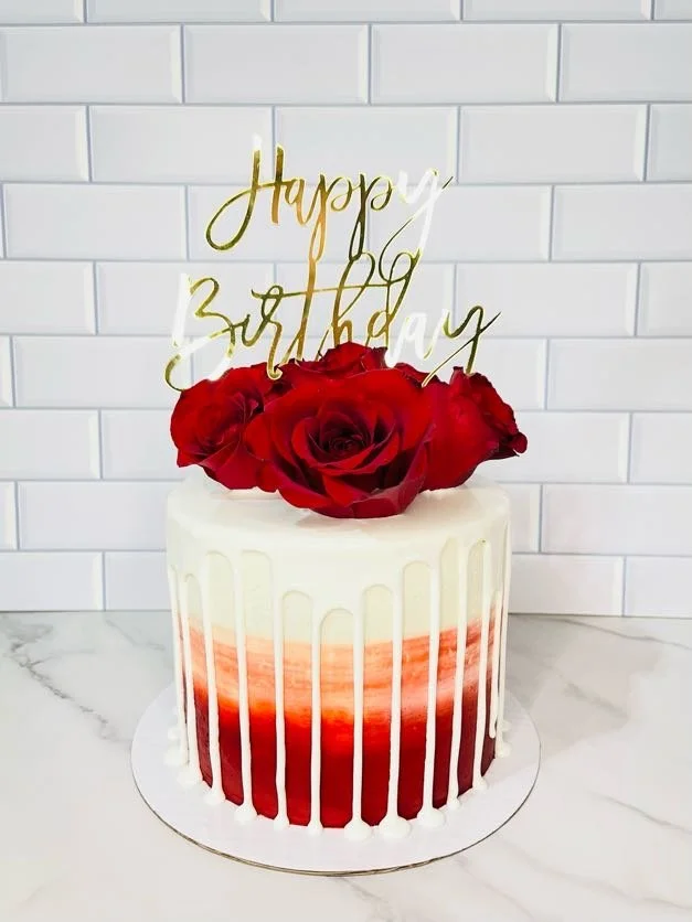 A birthday cake with white and red ombré frosting, decorated with red roses on top, and a gold 'Happy Birthday' cake topper, set against a tiled white background.