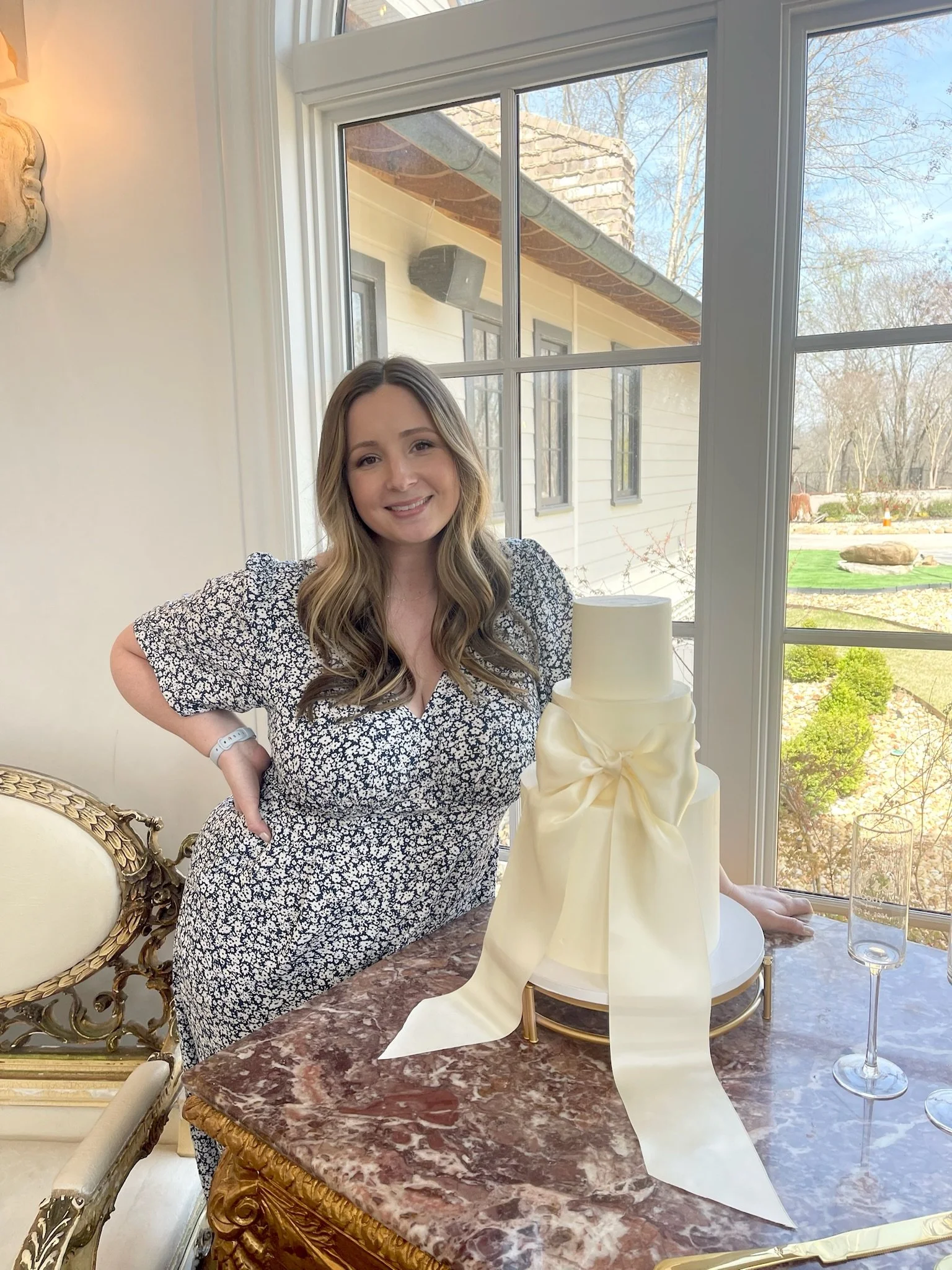 A young woman with wavy light brown hair smiling and posing next to a multi-tiered white cake with a large satin bow on a table in a bright room with large windows and a view of a garden outside.