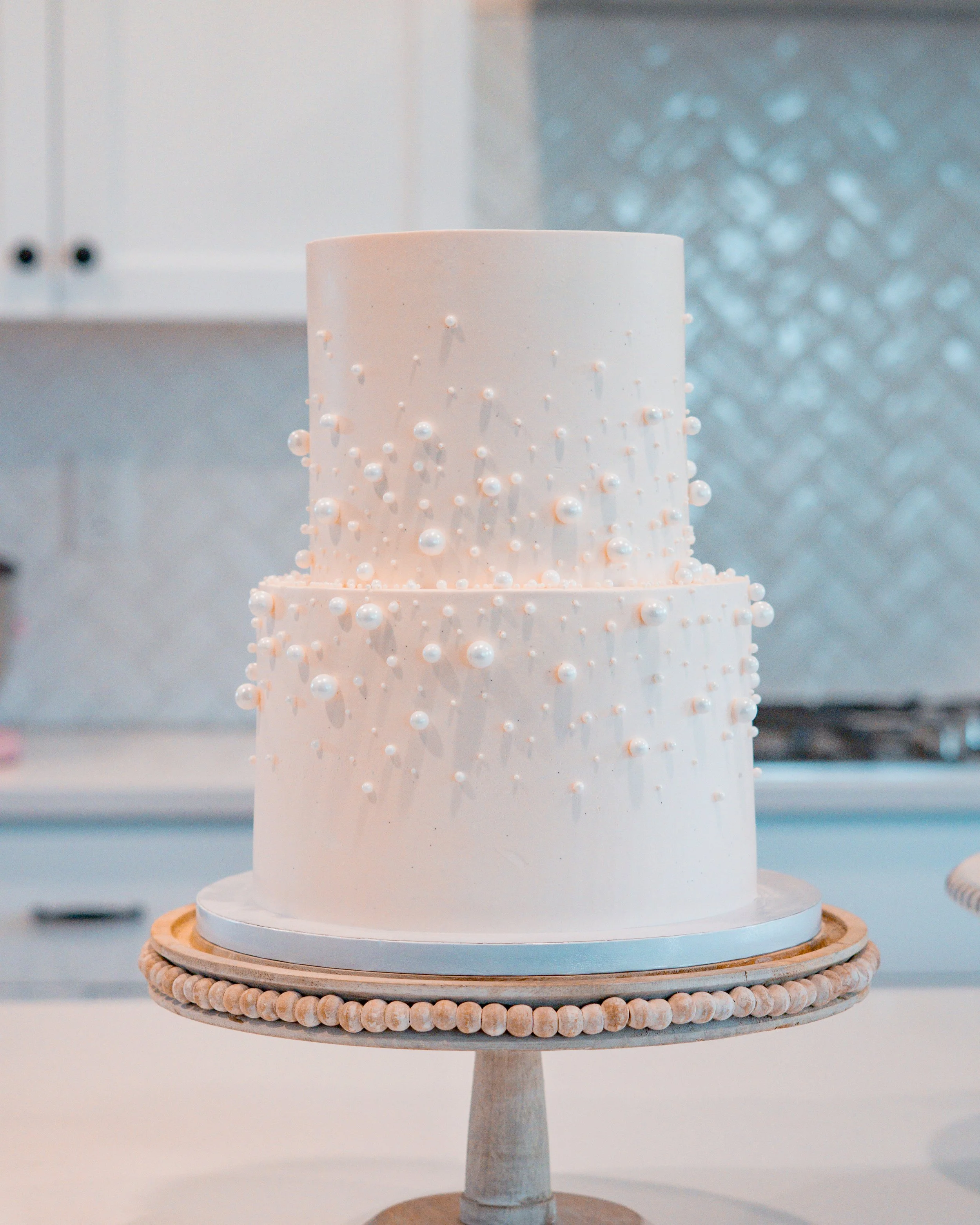 A white, cylindrical, multi-tiered cake decorated with small white pearl-like embellishments, displayed on a wooden cake stand with a decorative beaded border.