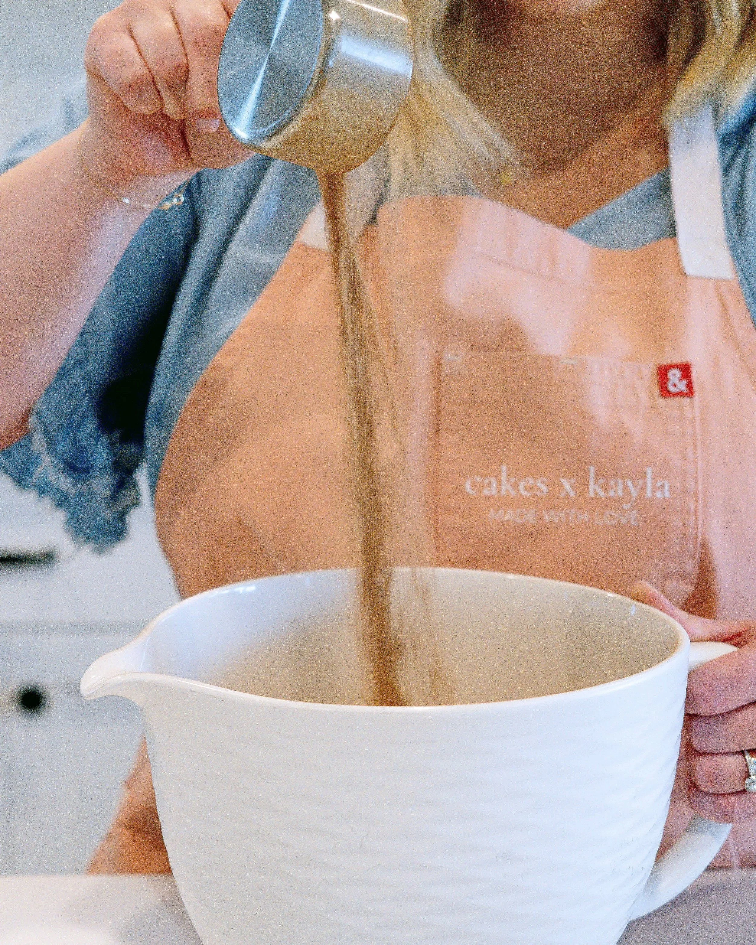 A person is pouring brown sugar from a metal container into a white mug, with a pink baking box in the background that has text reading 'cakes x kayla' and 'made with love'.