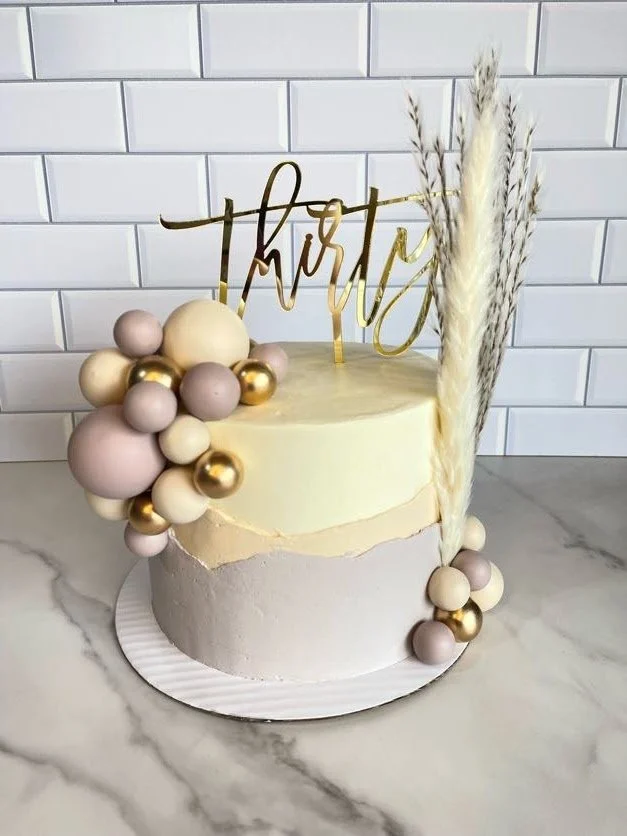 A two-tiered cake decorated with pastel-colored and gold spherical balloons, dried pampas grass, and a gold 'thirty' cake topper, placed on a white marble surface against a white subway tile wall.