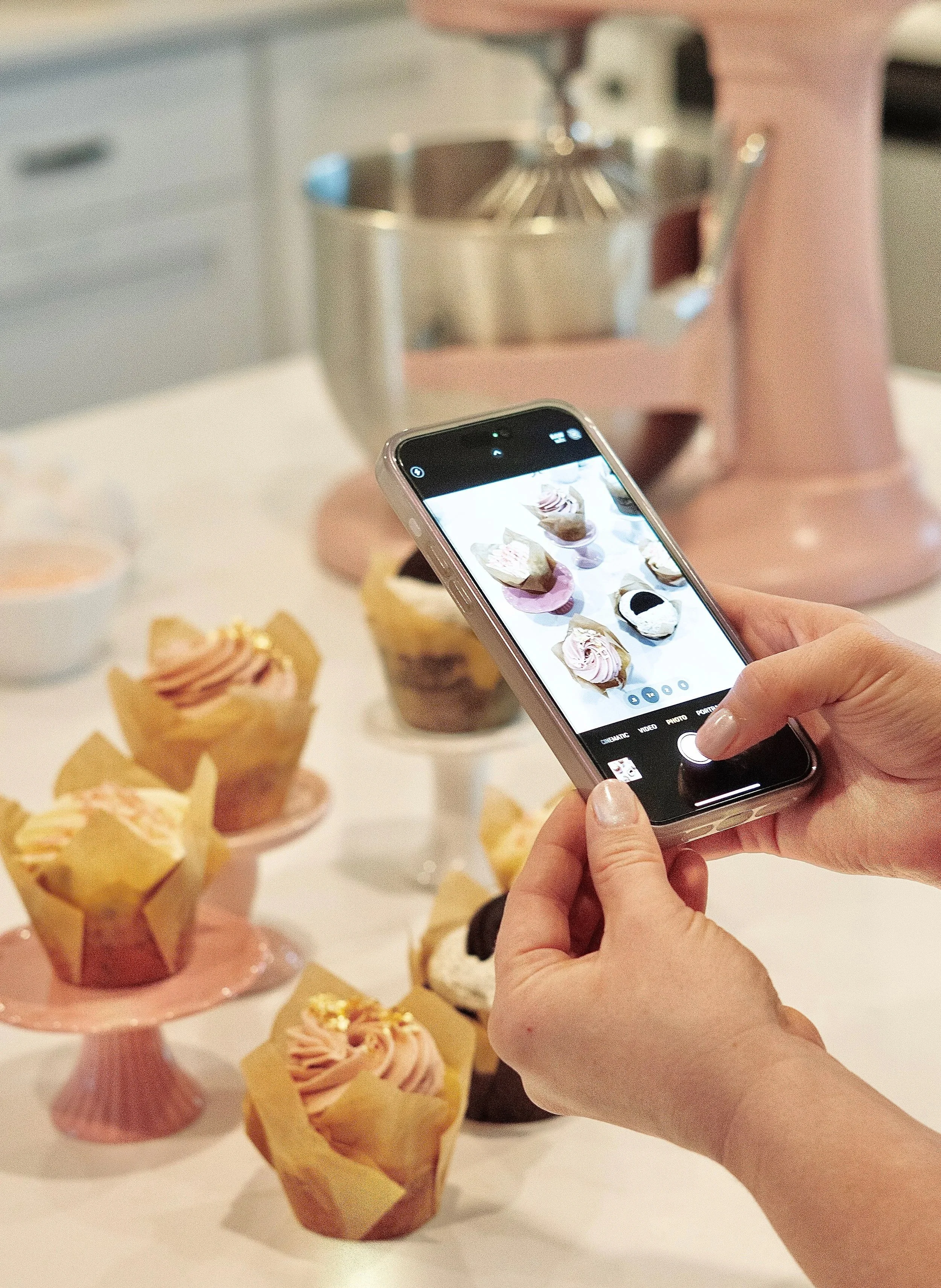Person taking a photo of cupcakes with a smartphone, cupcakes are decorated with pink, white, and black icing, displayed on a pink cake stand in the background.