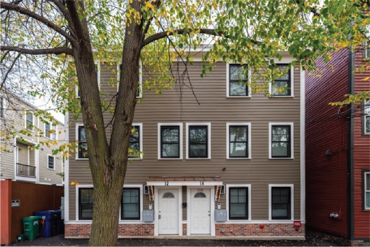 Three-story residential building with brown siding, multiple windows, two white front doors, and a large tree in the foreground.