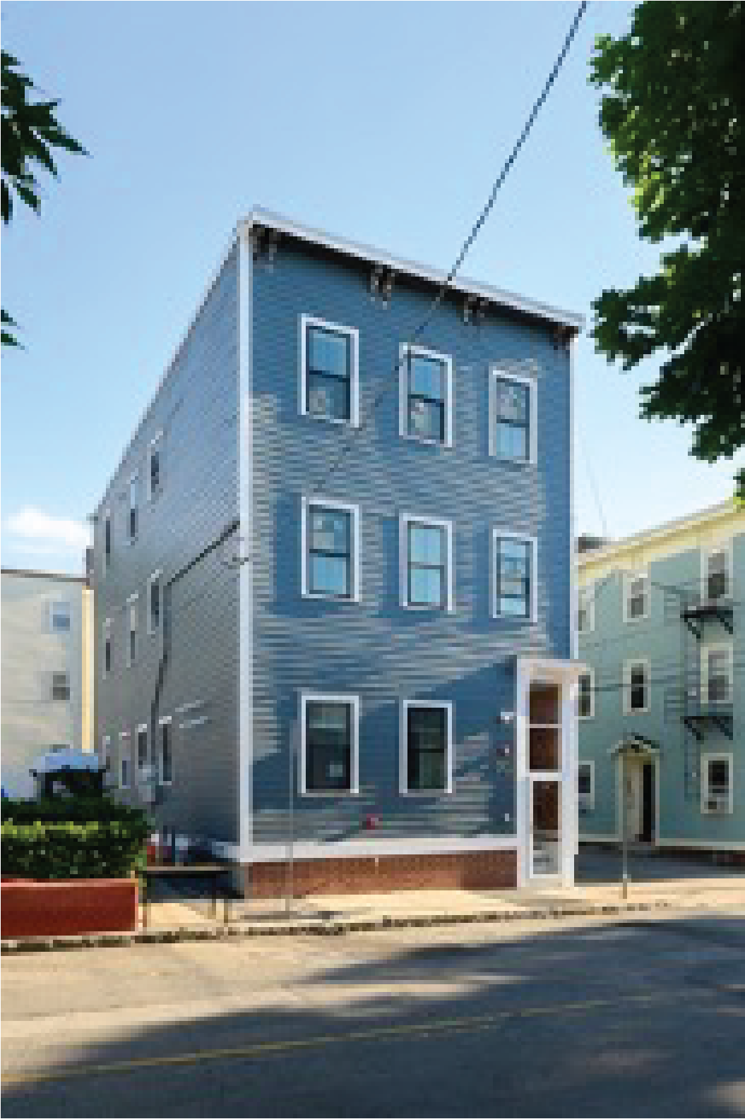 Three-story blue wooden building with white trim and multiple windows on a city street, surrounded by other similar residential buildings and trees.