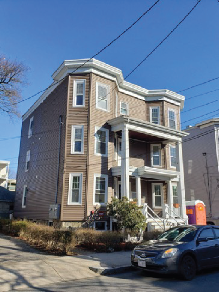 Three-story brown residential house with white trim, featuring a front porch and parked car, under a clear blue sky.