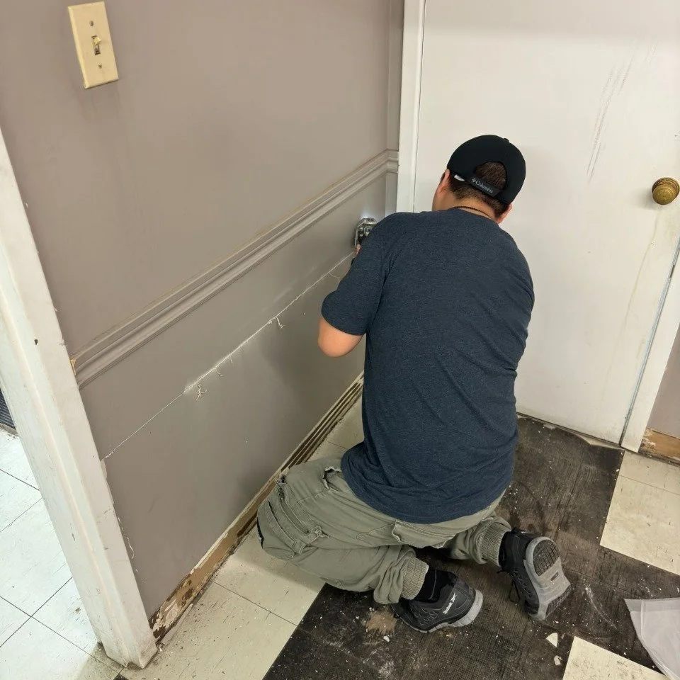Homem consertando drywall ao lado de uma porta em um interior, usando boné preto e camiseta cinza-escuro.