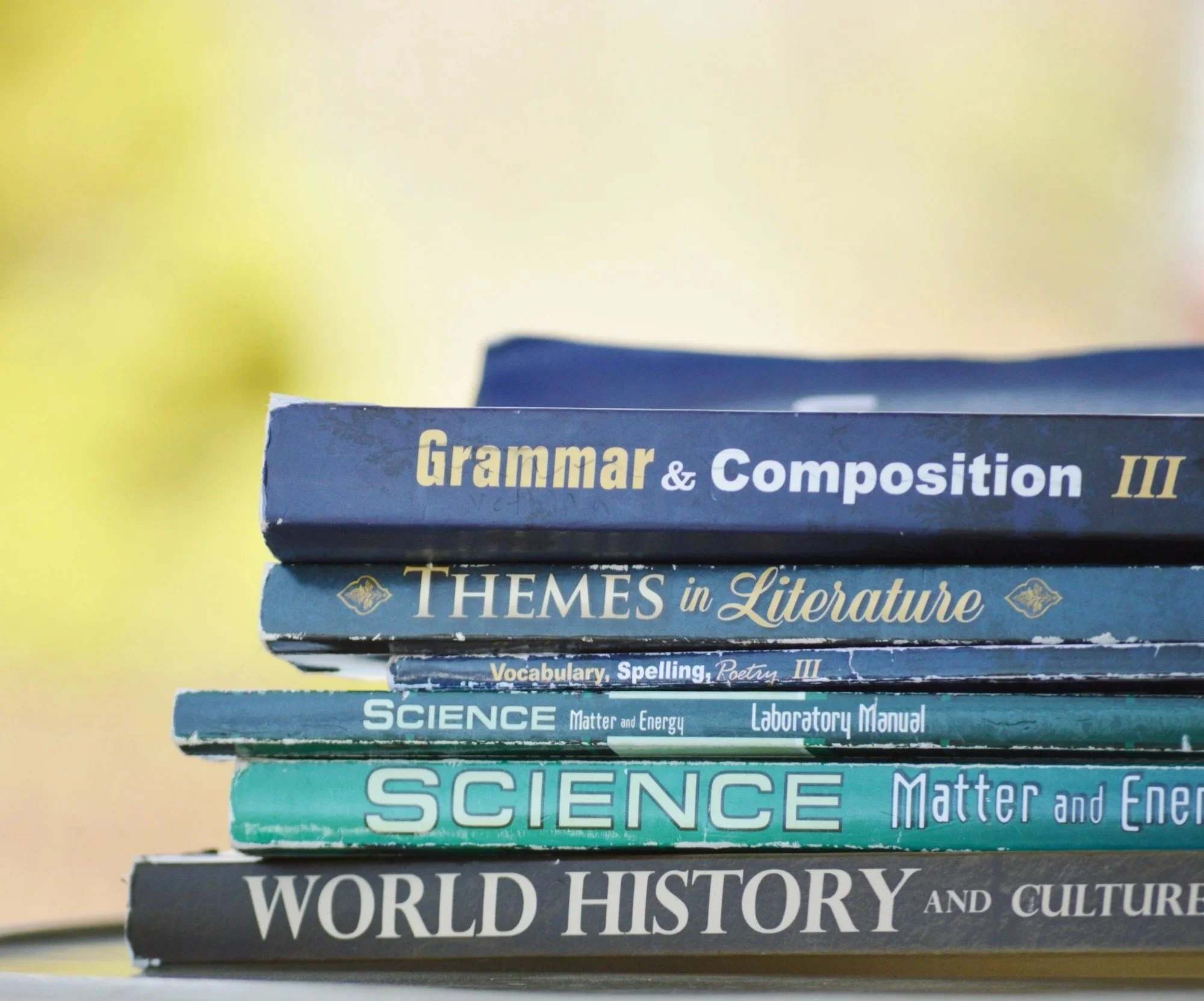 Stack of school textbooks on various subjects including Grammar & Composition, Themes in Literature, Vocabulary, Spelling, Poetry, Science, Matter and Energy, Laboratory Manual, World History, and Culture.