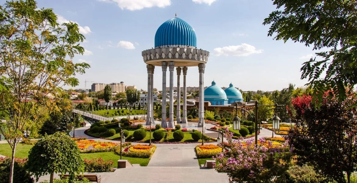A park with a decorative blue-domed pavilion on tall columns, surrounded by colorful flower beds, greenery, and trees under a clear sky.