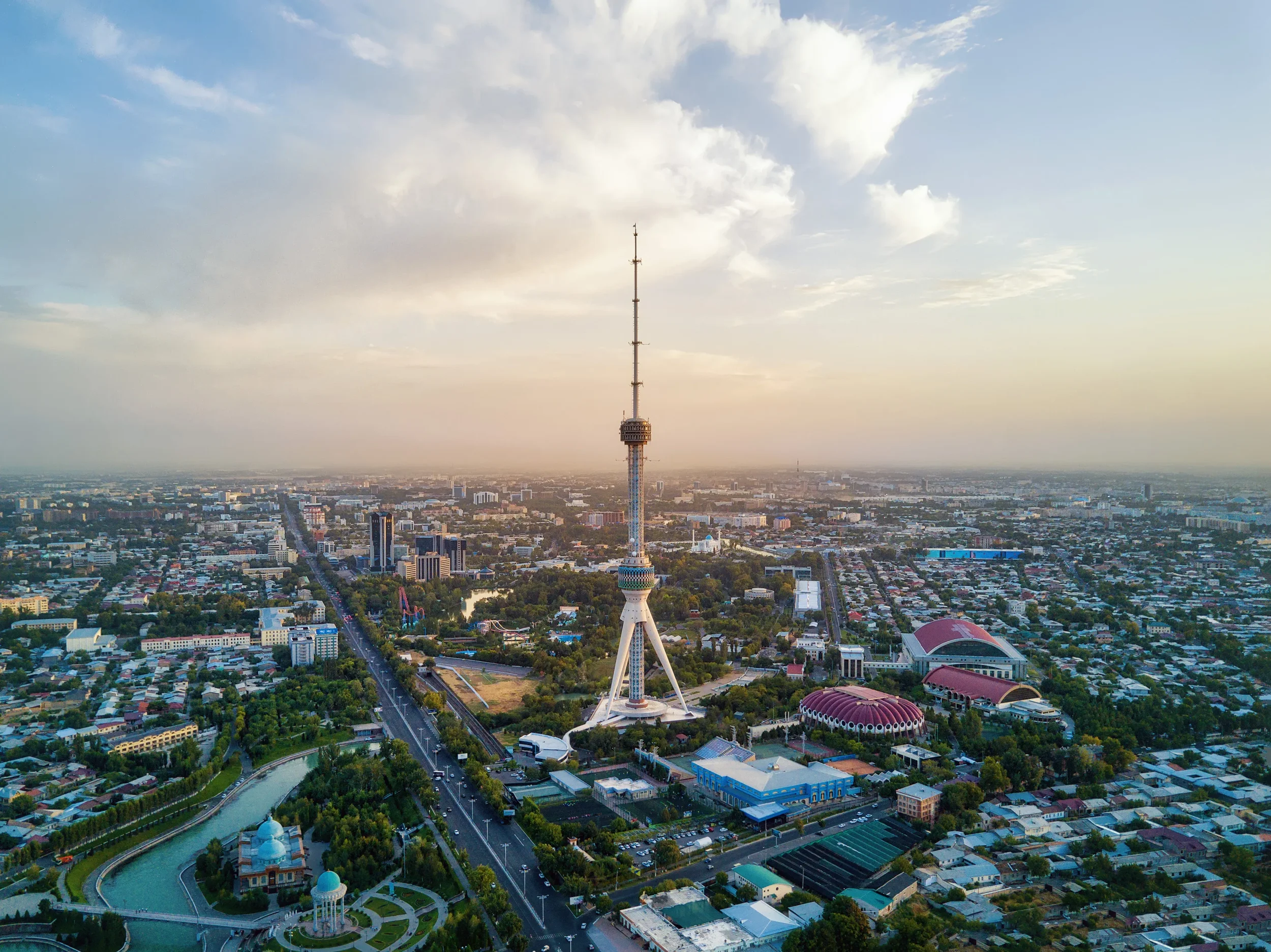 Aerial view of a city with a tall tower at the center, surrounding buildings, parks, and roads, under a partly cloudy sky during sunset.