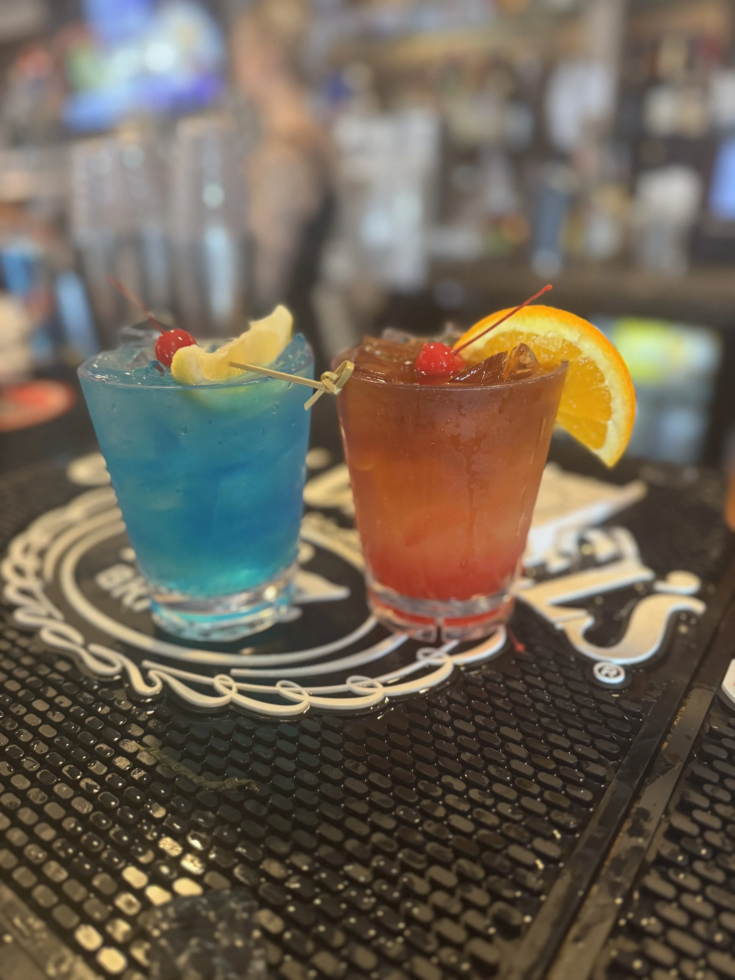 Two colorful cocktails, one blue and one red-orange, garnished with cherries and citrus slices, on a bar counter with a blurred bar background.