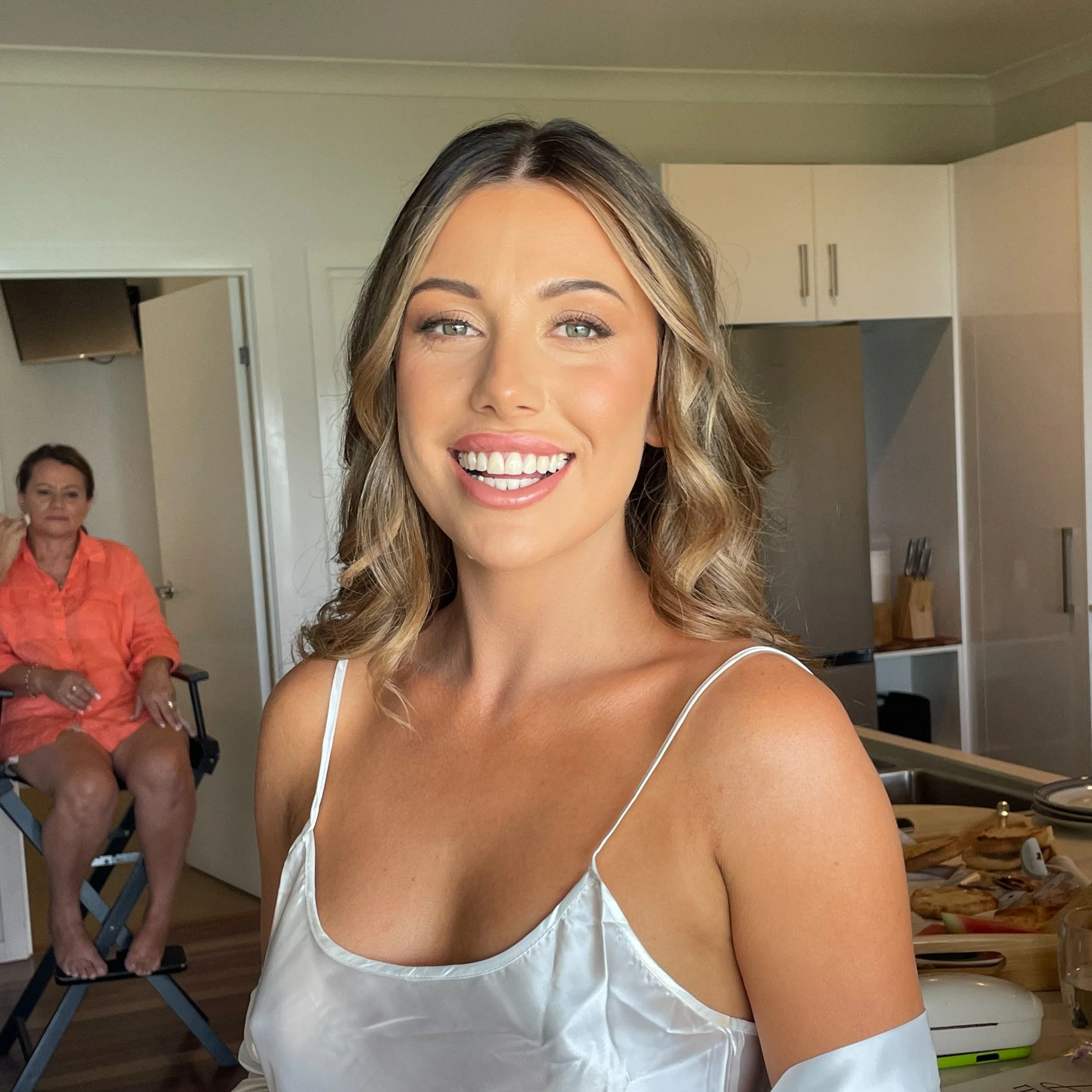 Young woman with wavy hair and light top smiling in a kitchen, with an older woman sitting in the background.
