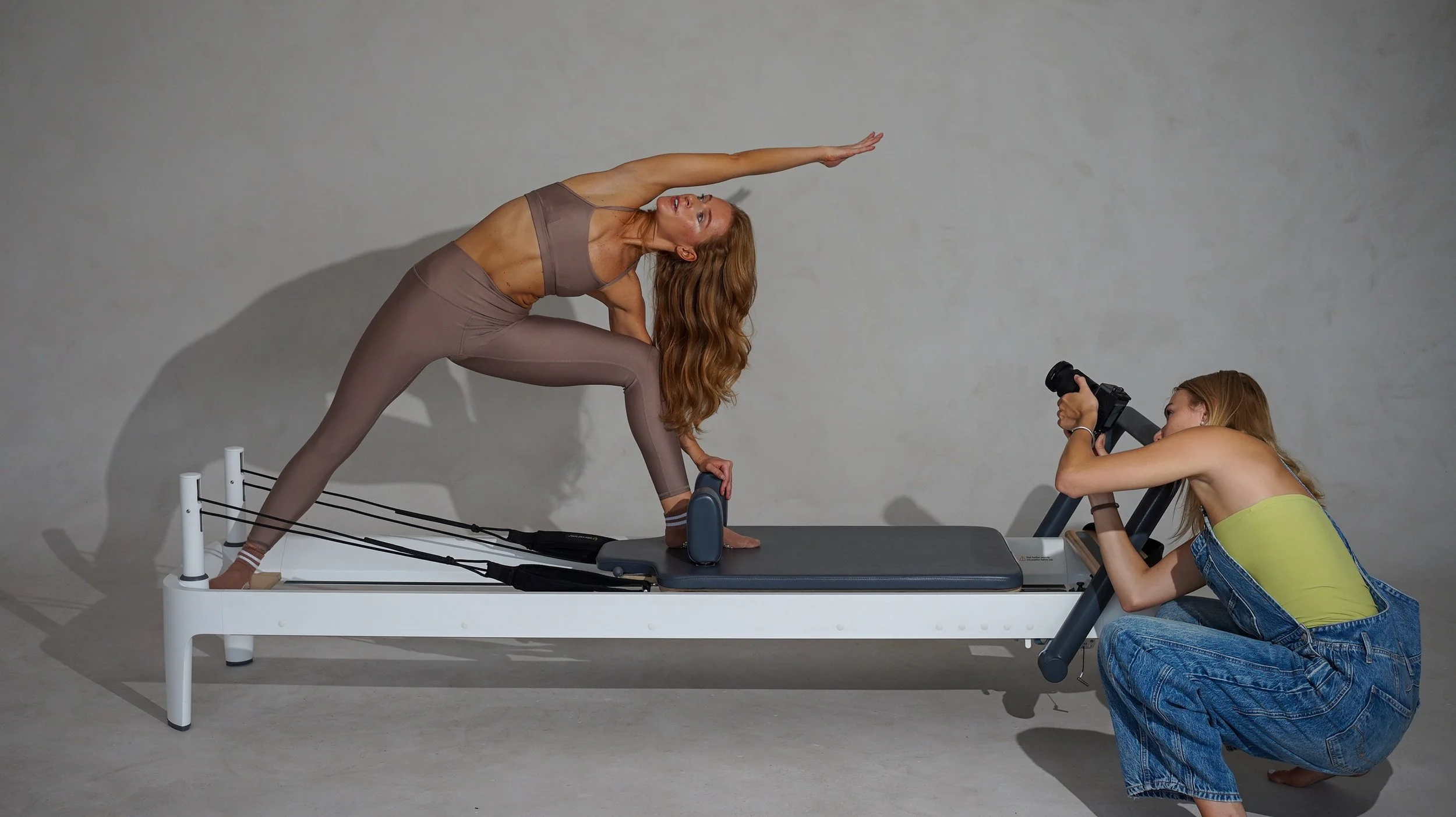 A woman practicing yoga on a reformer machine, captured with a camera held by a woman in overalls, in a studio with a plain wall background.
