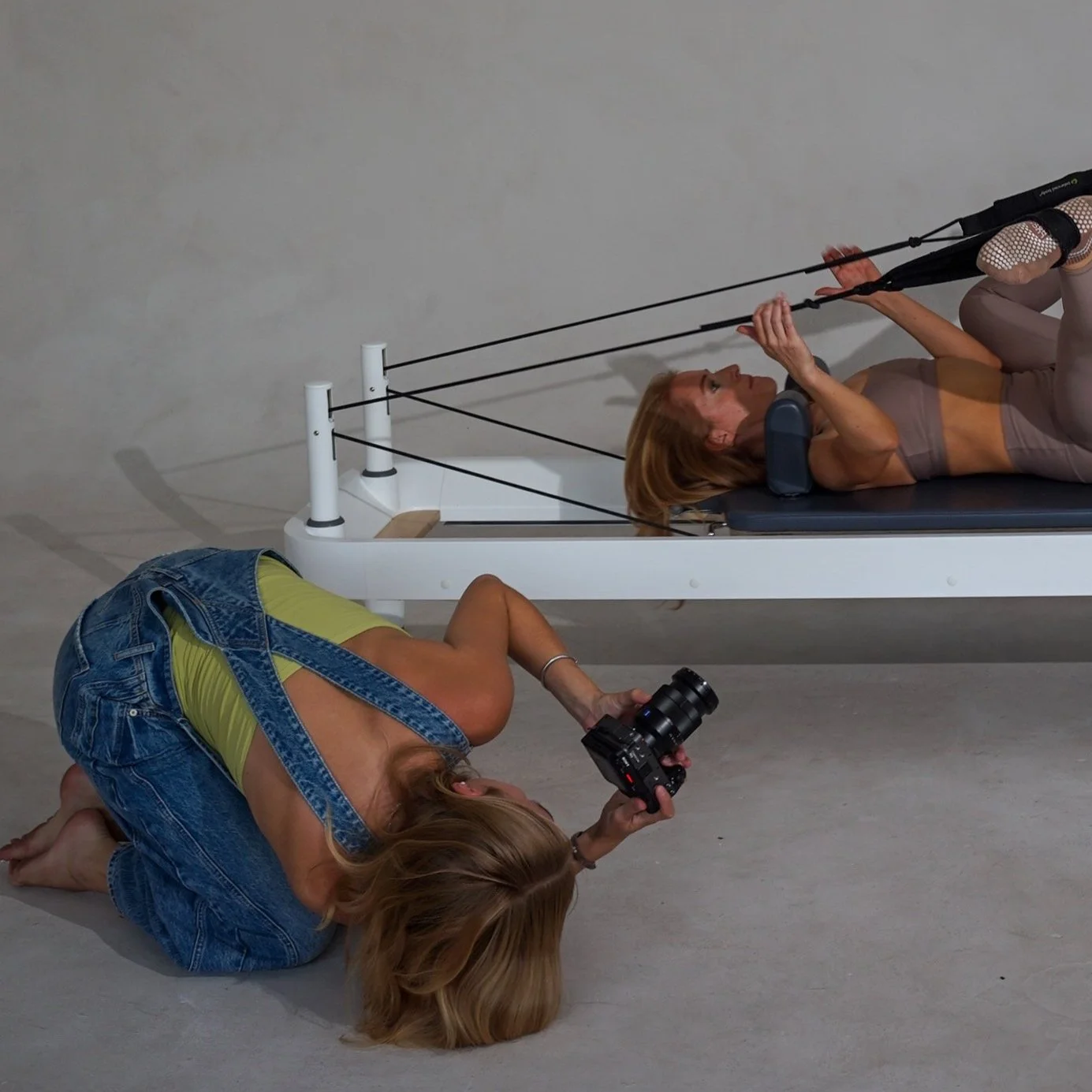 A woman lying on a Pilates reformer machine during an exercise session, with a photographer kneeling on the floor taking her picture from a low angle.