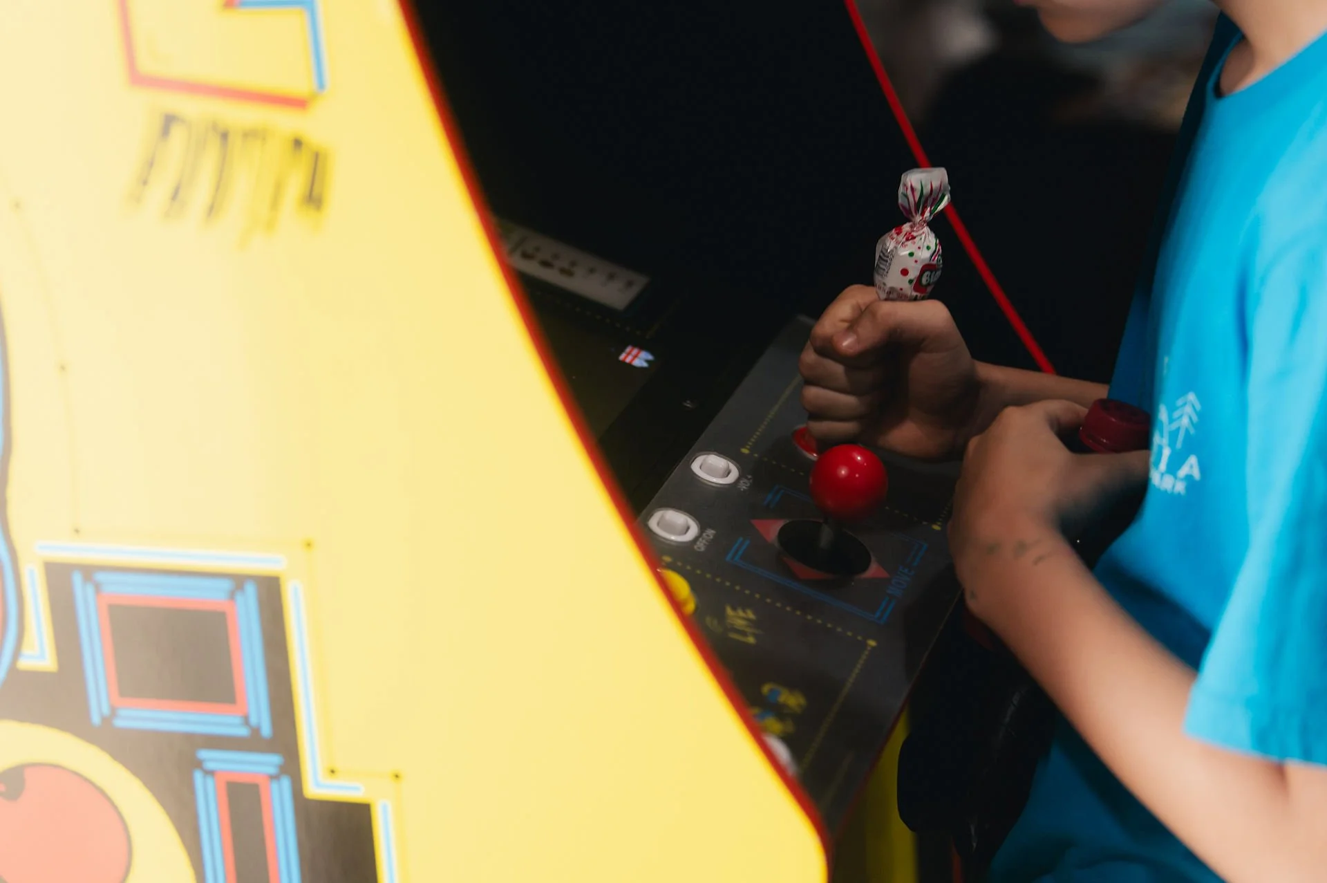 Close-up of a person playing an arcade game, holding a lever with a red ball at the bottom and a small wrapped candy in their other hand, partially visible. The arcade game panel has yellow and black parts with some buttons and a screen.