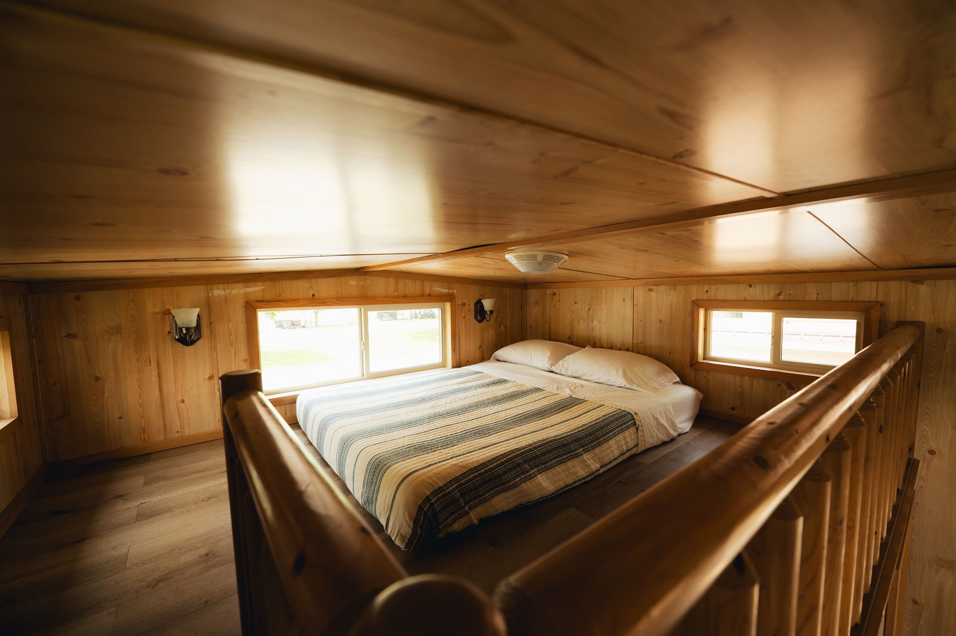 Wooden-finished loft bedroom with a bed, two pillows, striped bedspread, small windows, and wall-mounted light fixtures.