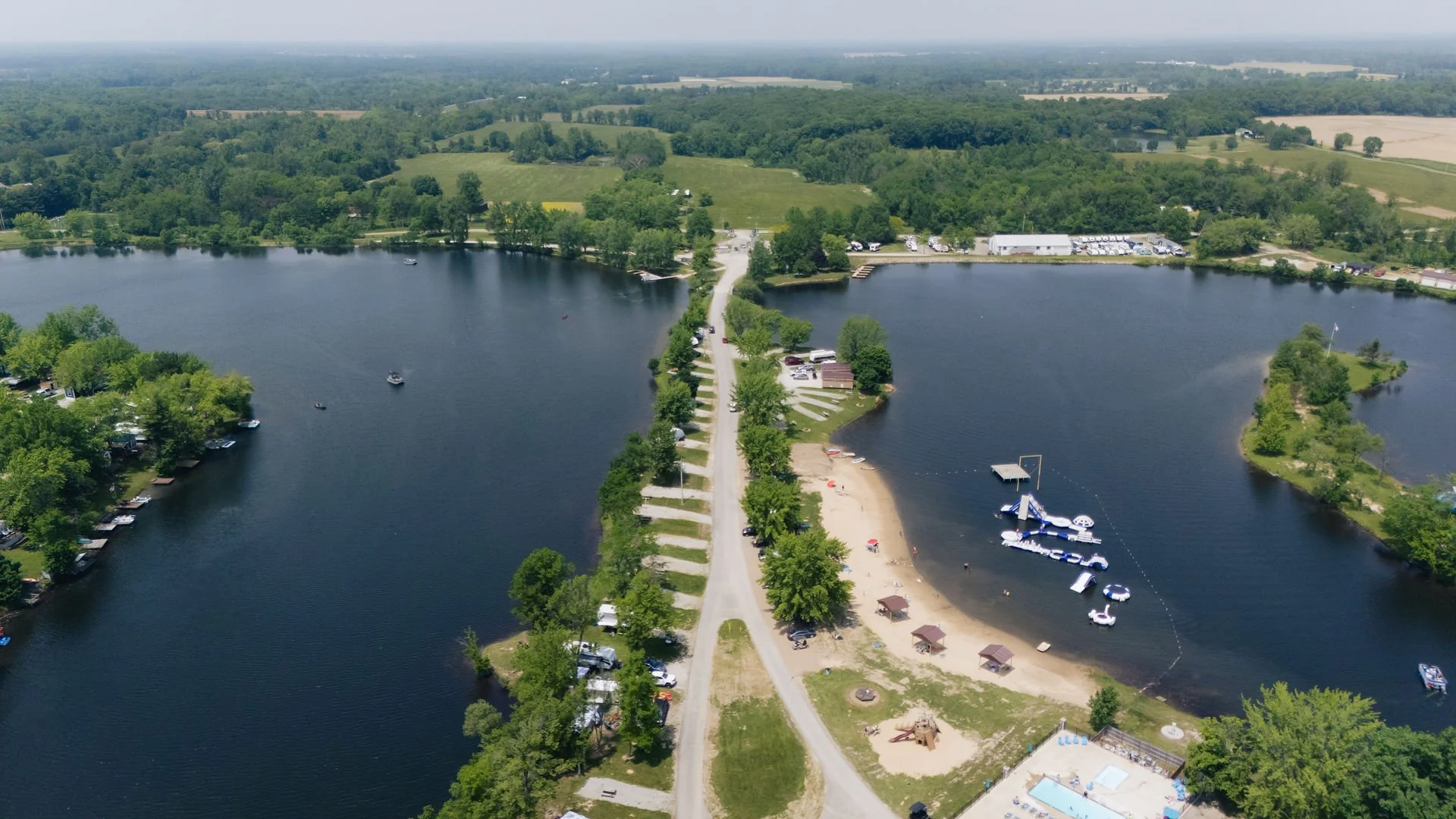 Aerial view of a lakeside park with boats, a floating slide, and a sandy beach area, surrounded by green trees and open fields in the background.