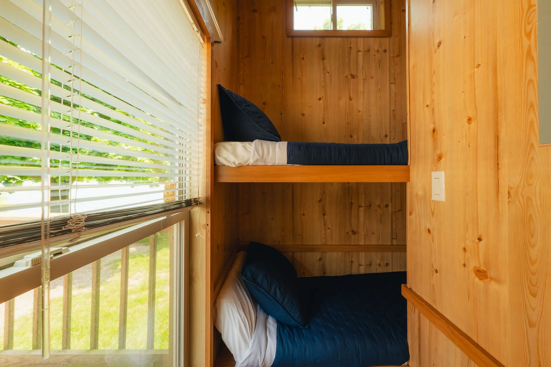 A wooden cabin room with two beds, one above the other. The beds have blue blankets and pillows. There are large windows with white blinds and a small window near the ceiling, allowing natural light into the room.
