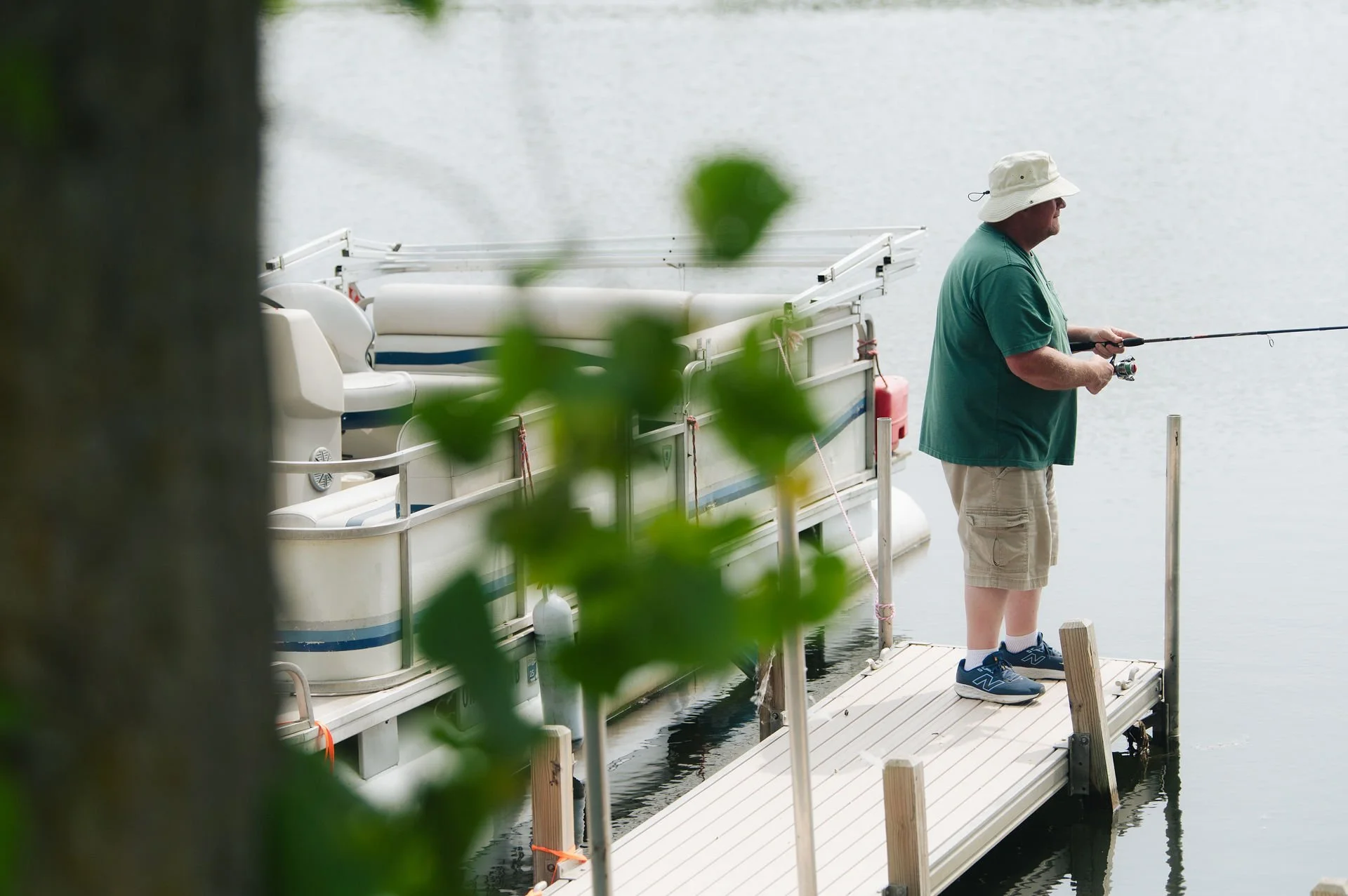 A man standing on a wooden dock fishing in a body of water, with a pontoon boat in the background.