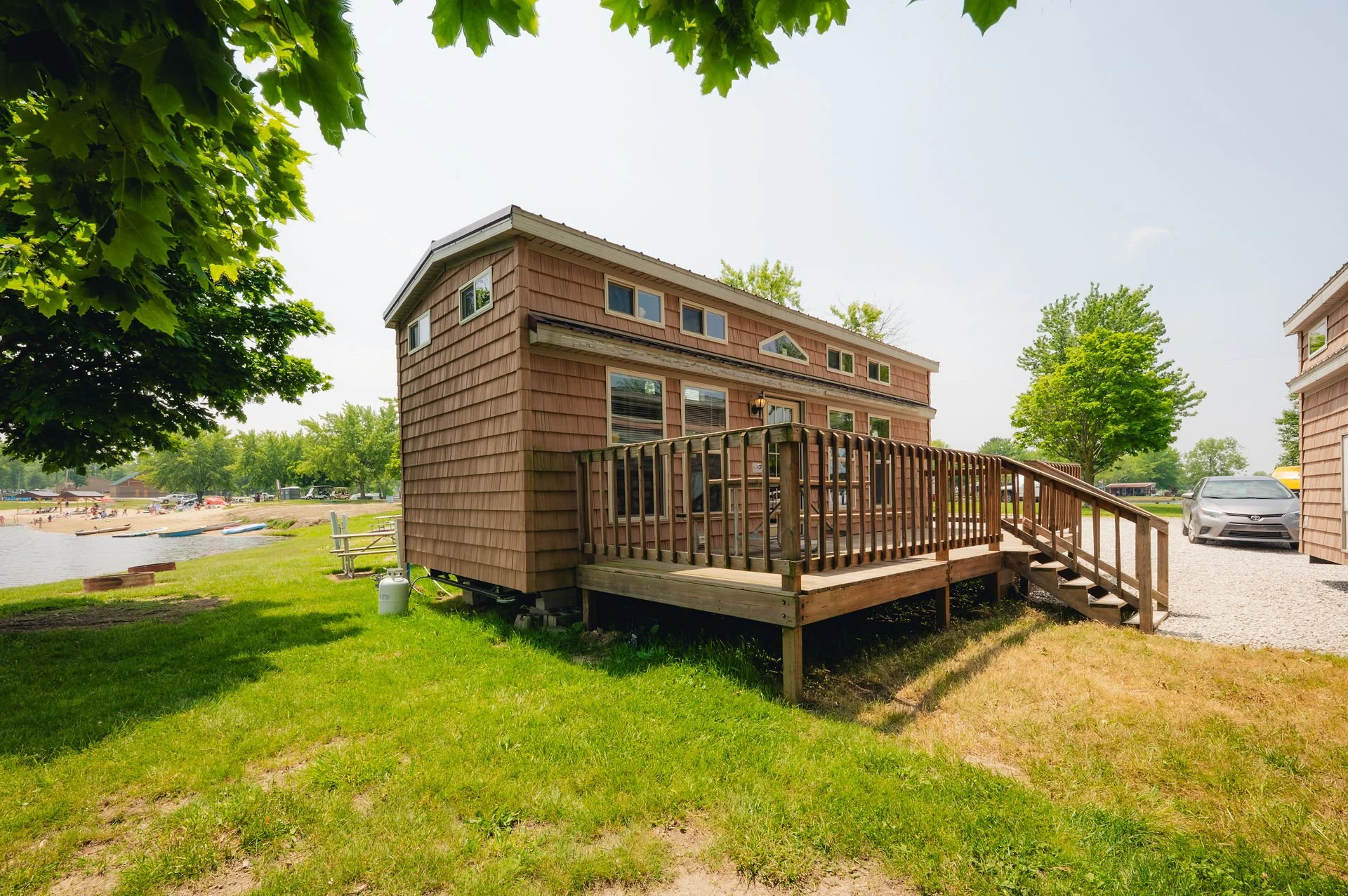 Brown house with a wooden deck overlooking a green lawn and a lake with people and paddle boats in the background.