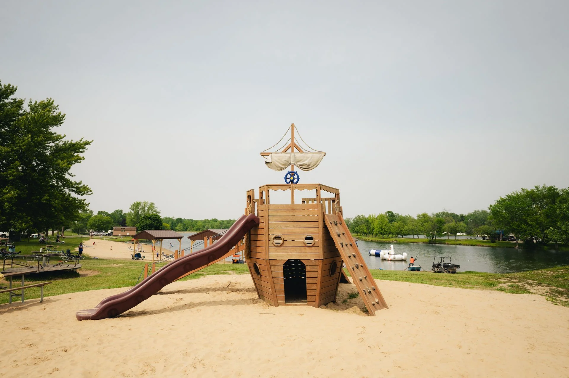 A wooden pirate ship-shaped playground with a slide and climbing wall on sandy ground near a lake, with trees and boats in the background.