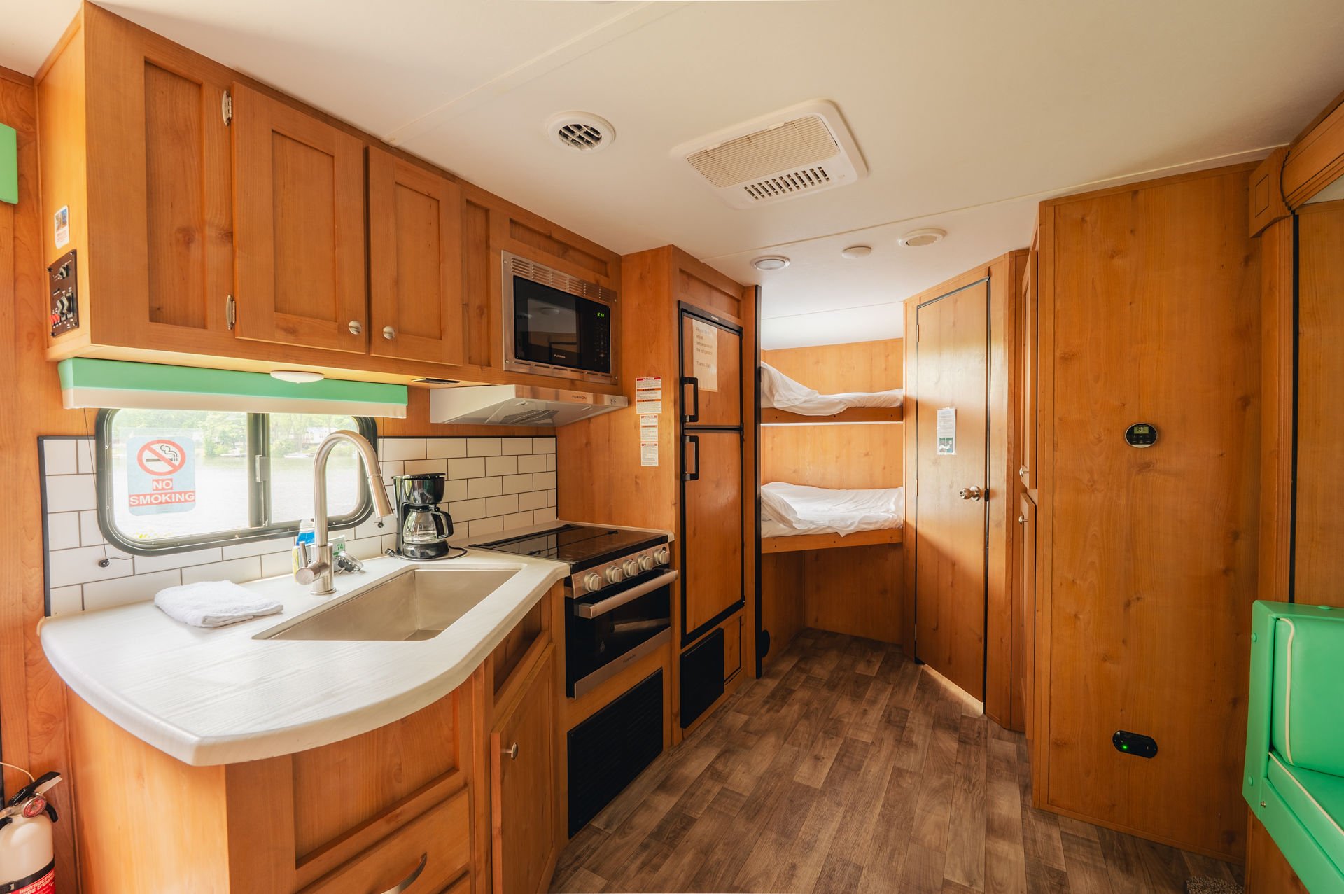 Interior of a small wooden cabin with a kitchenette, including a sink, coffee maker, microwave, stove, and refrigerator, with a set of wooden bunk beds in the background.