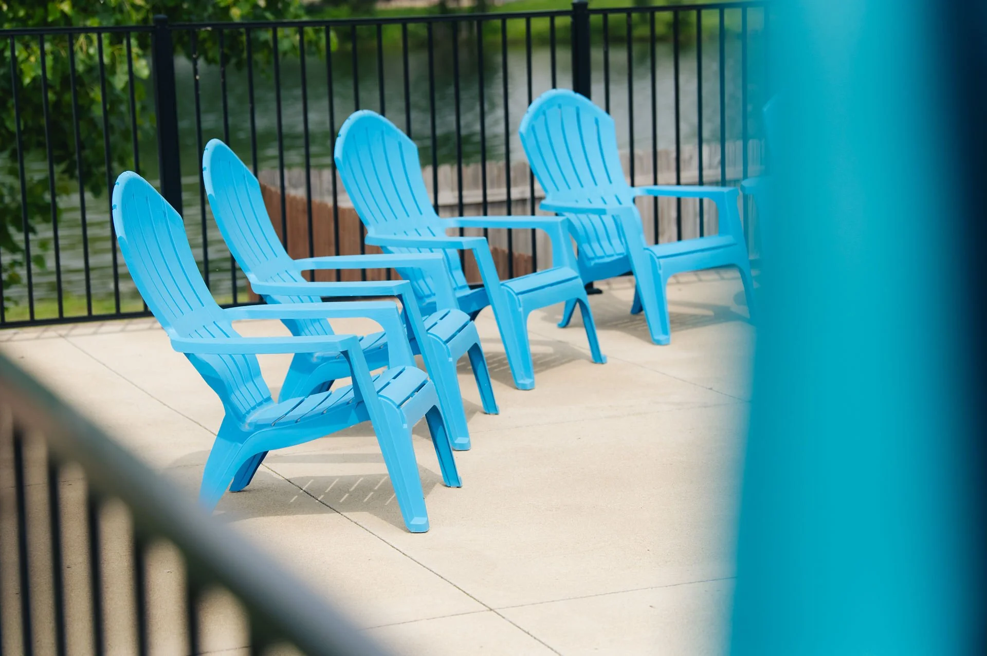 Four empty blue Adirondack chairs on a patio near a black metal fence with water and greenery in the background.