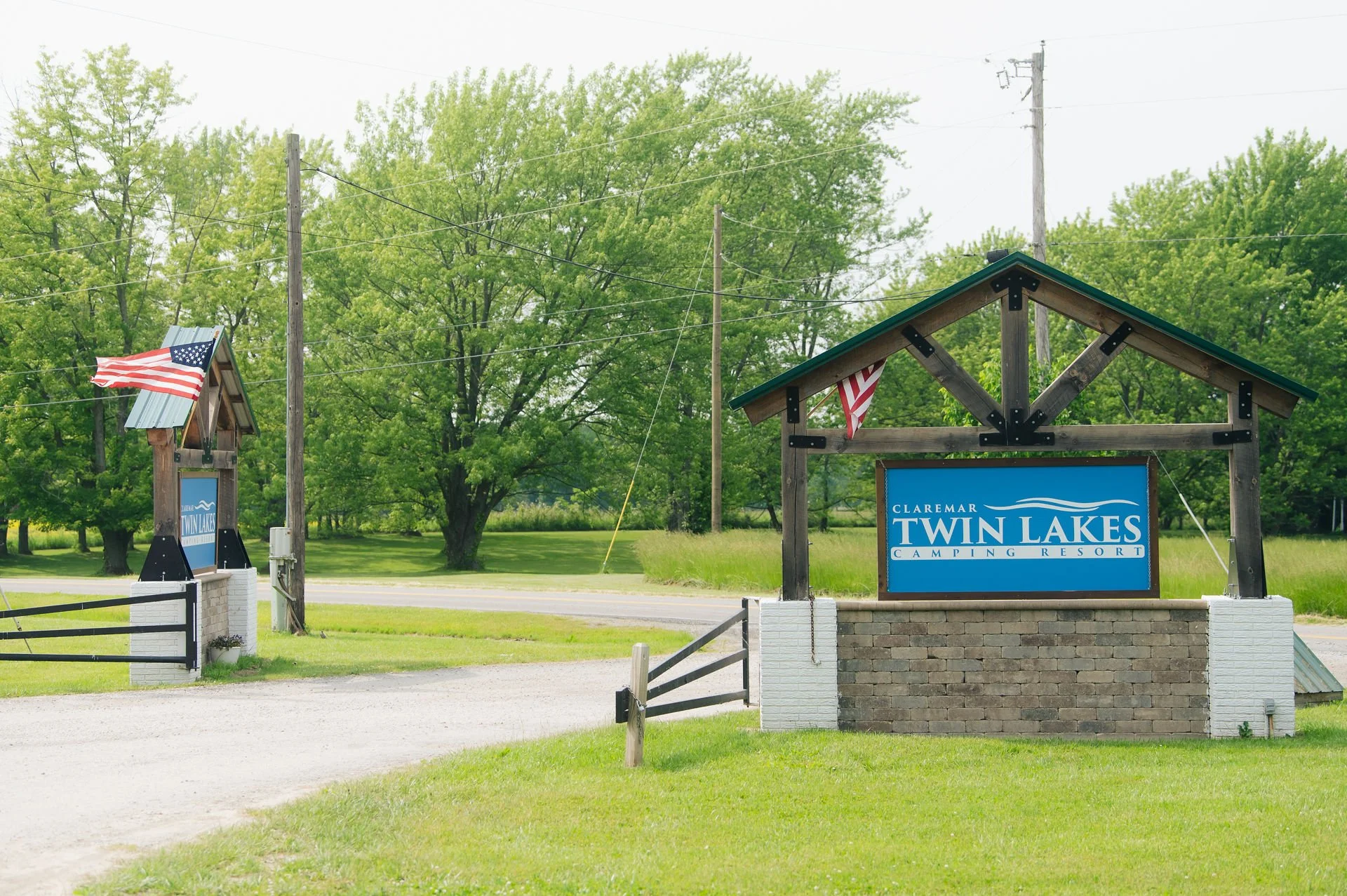 Entrance sign for Claremont Twin Lakes Camping Resort with American flags, trees, and a road in the background.
