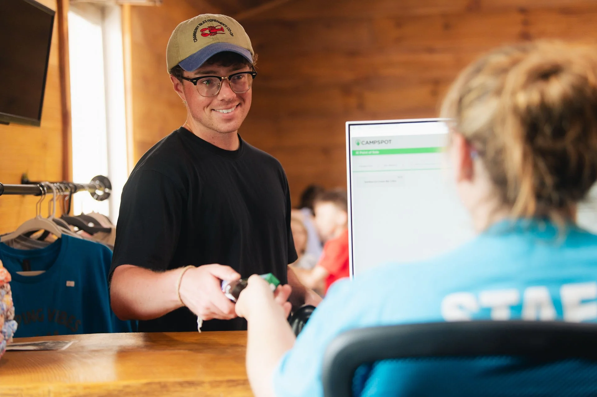 A young man is smiling while checking out at a reception desk, wearing glasses, a beige cap, and a black T-shirt, with a staff member assisting him.