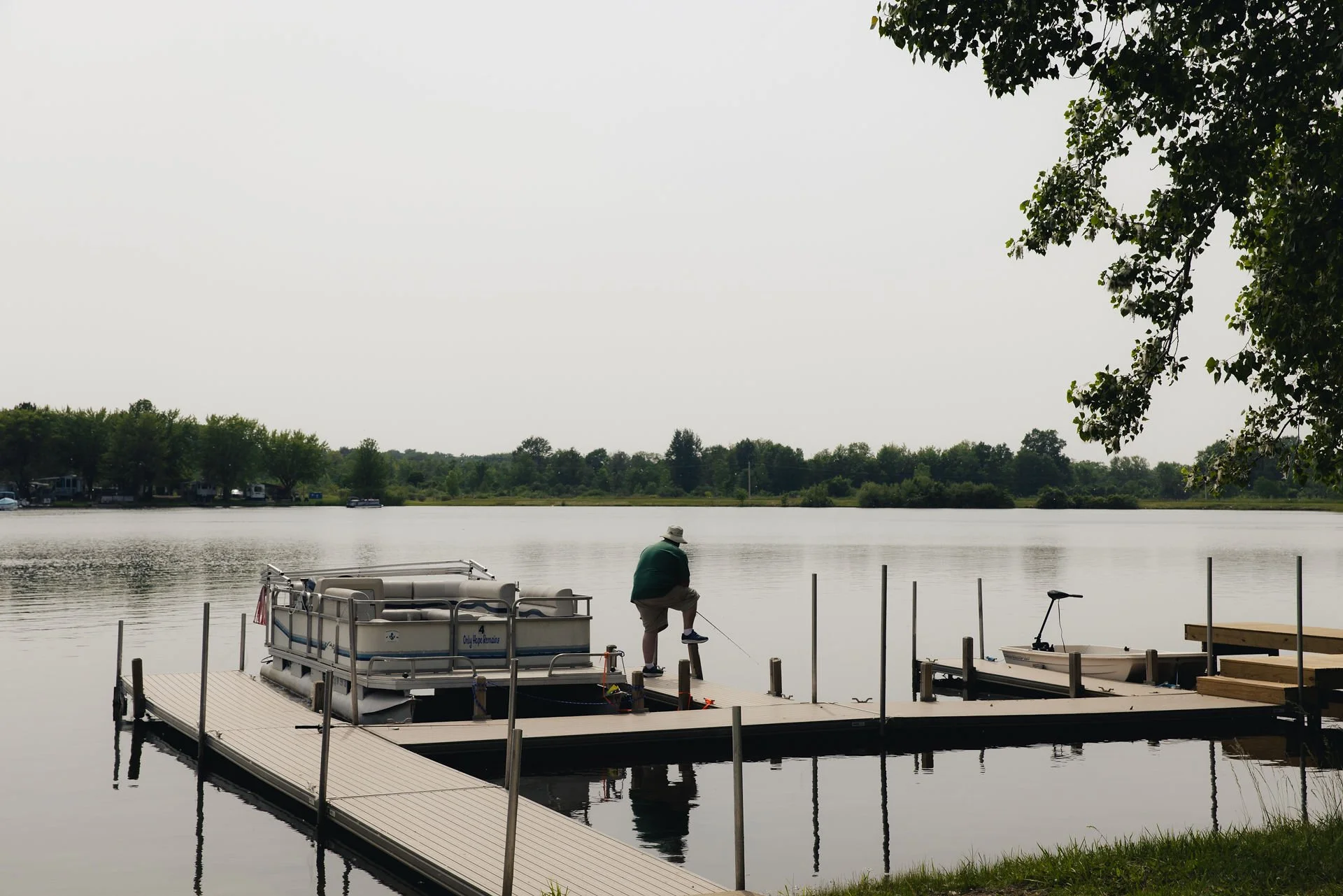 Man on a dock by a lake, sitting on a post with one foot lifted, with pontoon boat, a small boat with an outboard motor, trees, and calm water in the background.