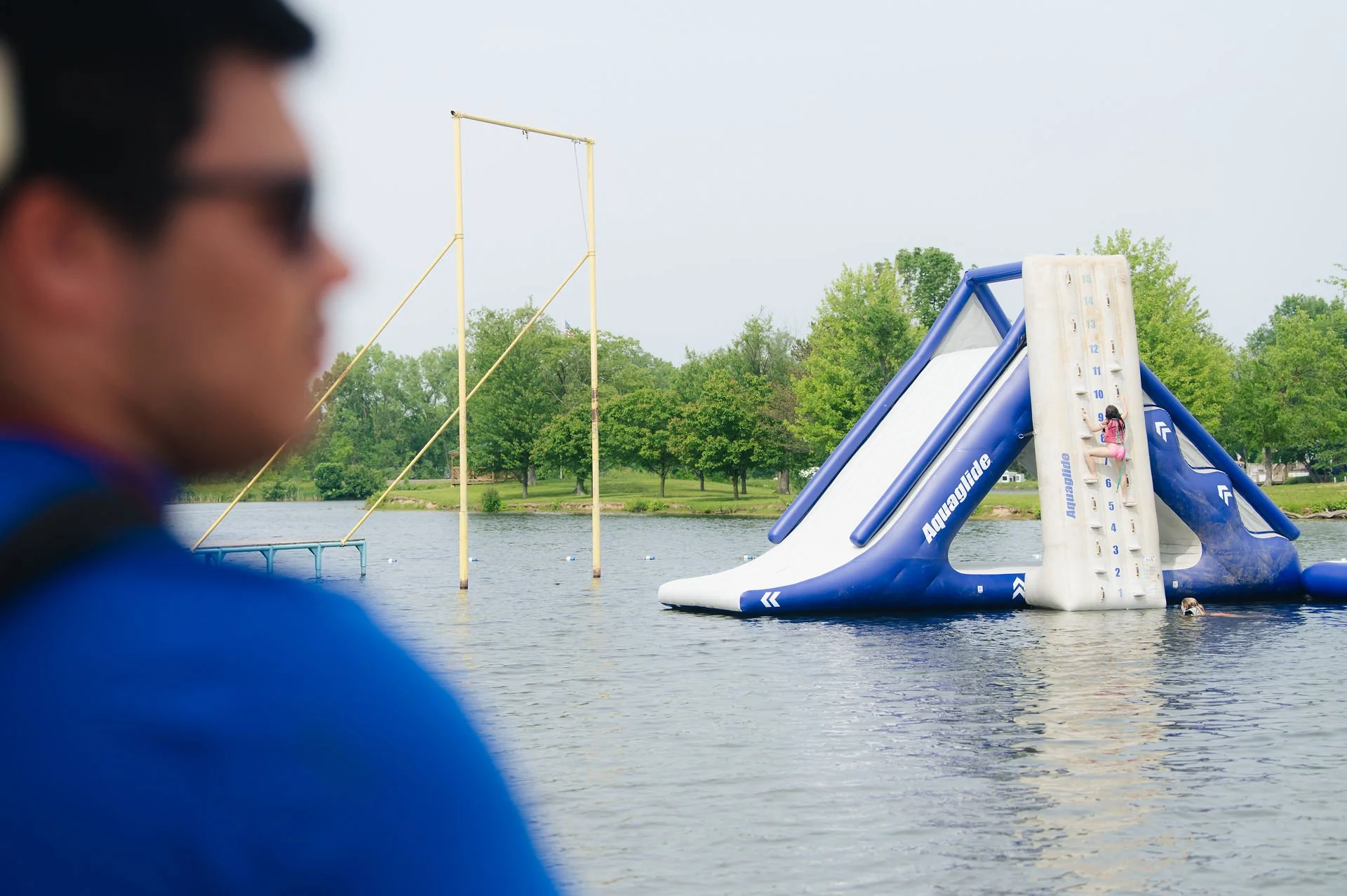 A person wearing sunglasses and a blue shirt is blurred in the foreground, with a large inflatable water slide and a high climbing wall attached to it in the water. The slide and climbing wall are part of an inflatable water park by a lake or river, 