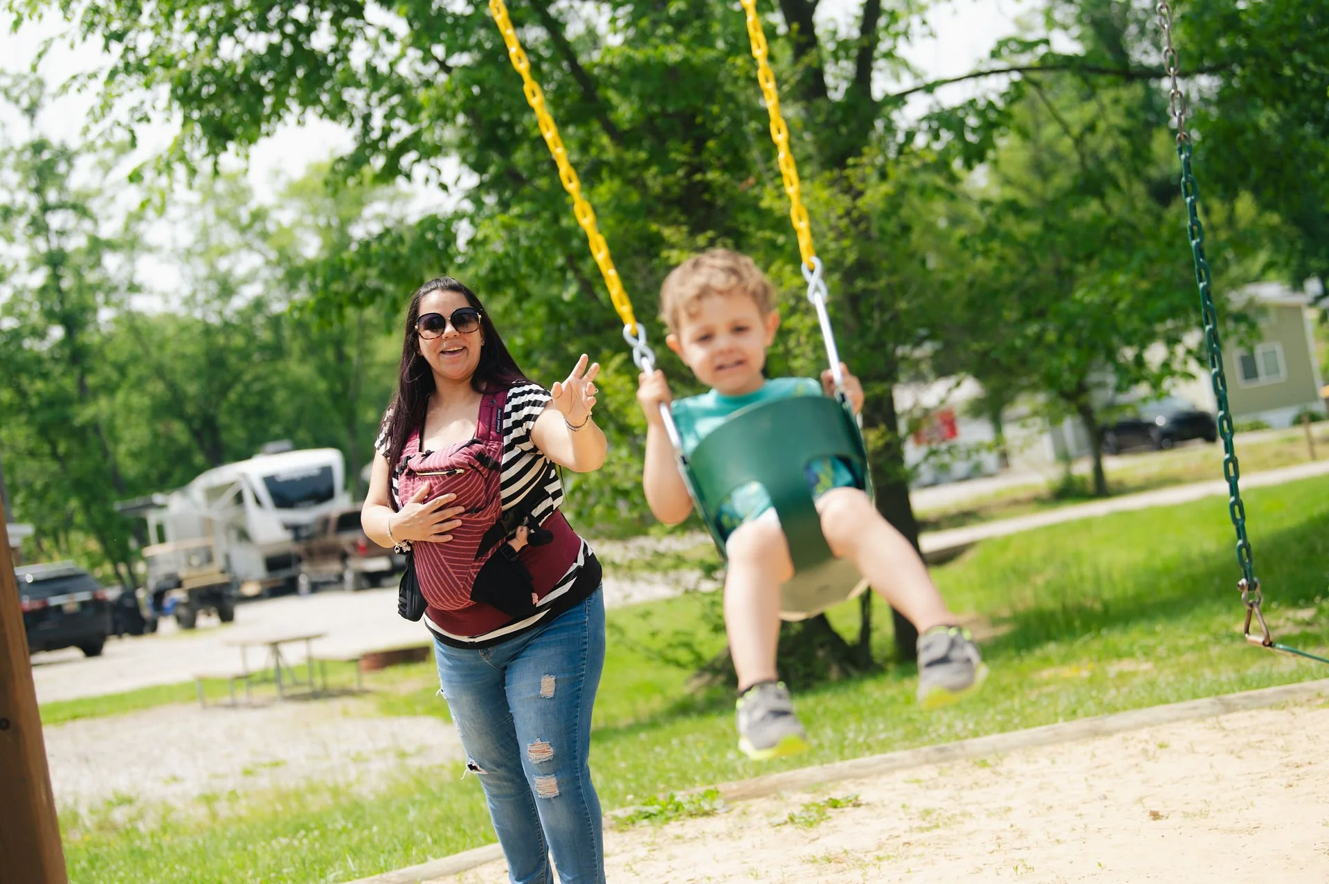 A woman and a young boy on a swing set in a park. The woman is smiling and pointing at the camera, while the boy is sitting in a green swing. The park has green grass, trees, and cars in the background.