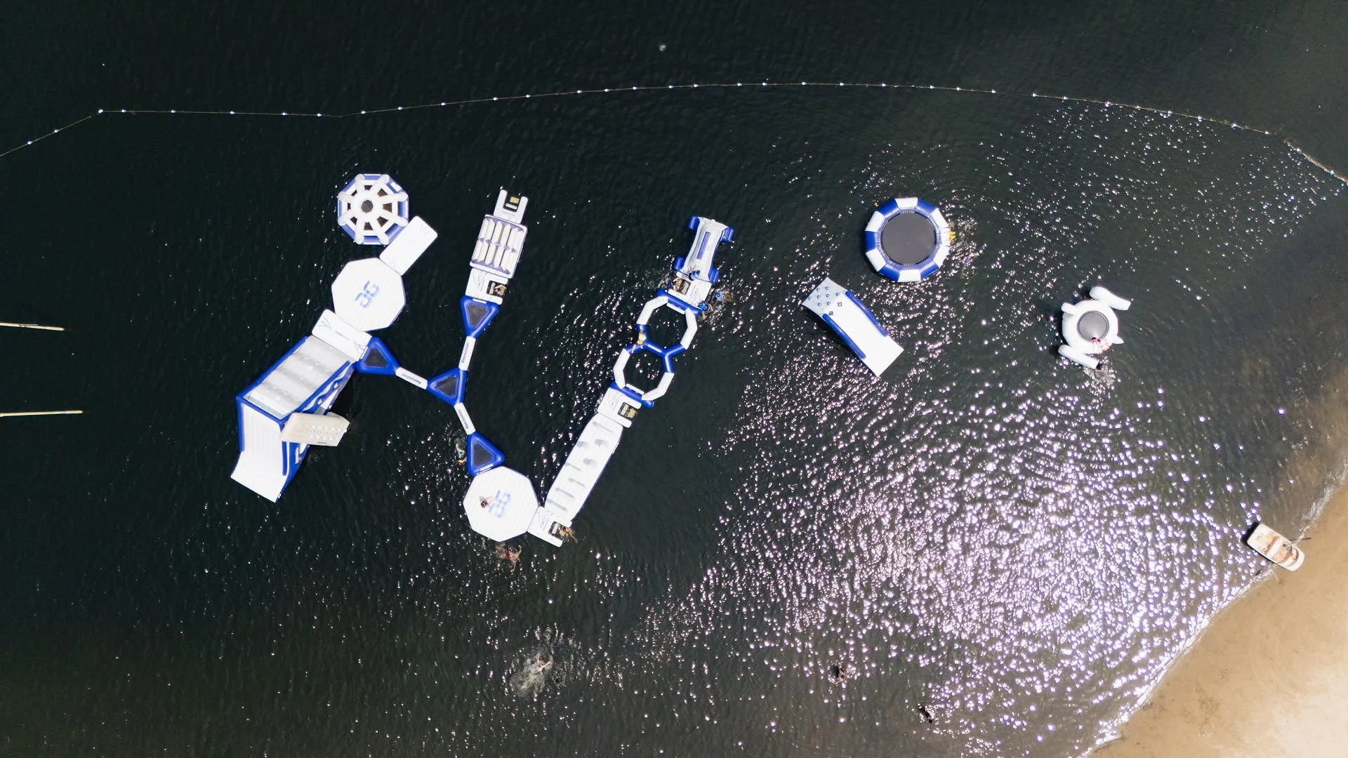 Aerial view of various water park floats and structures arranged on a body of water with a sandy beach on the right side.