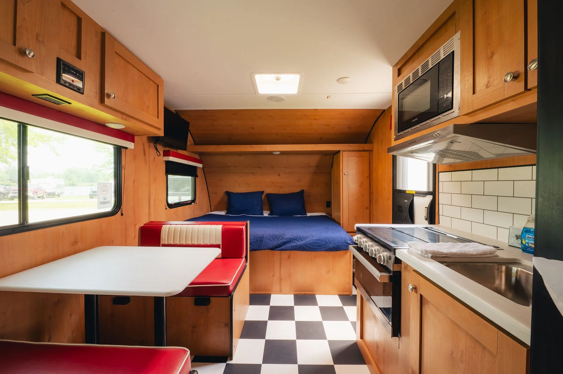 Interior of a compact camper or RV with a small kitchen area, dining table with red and white seats, and a sleeping area with a bed and blue bedding.