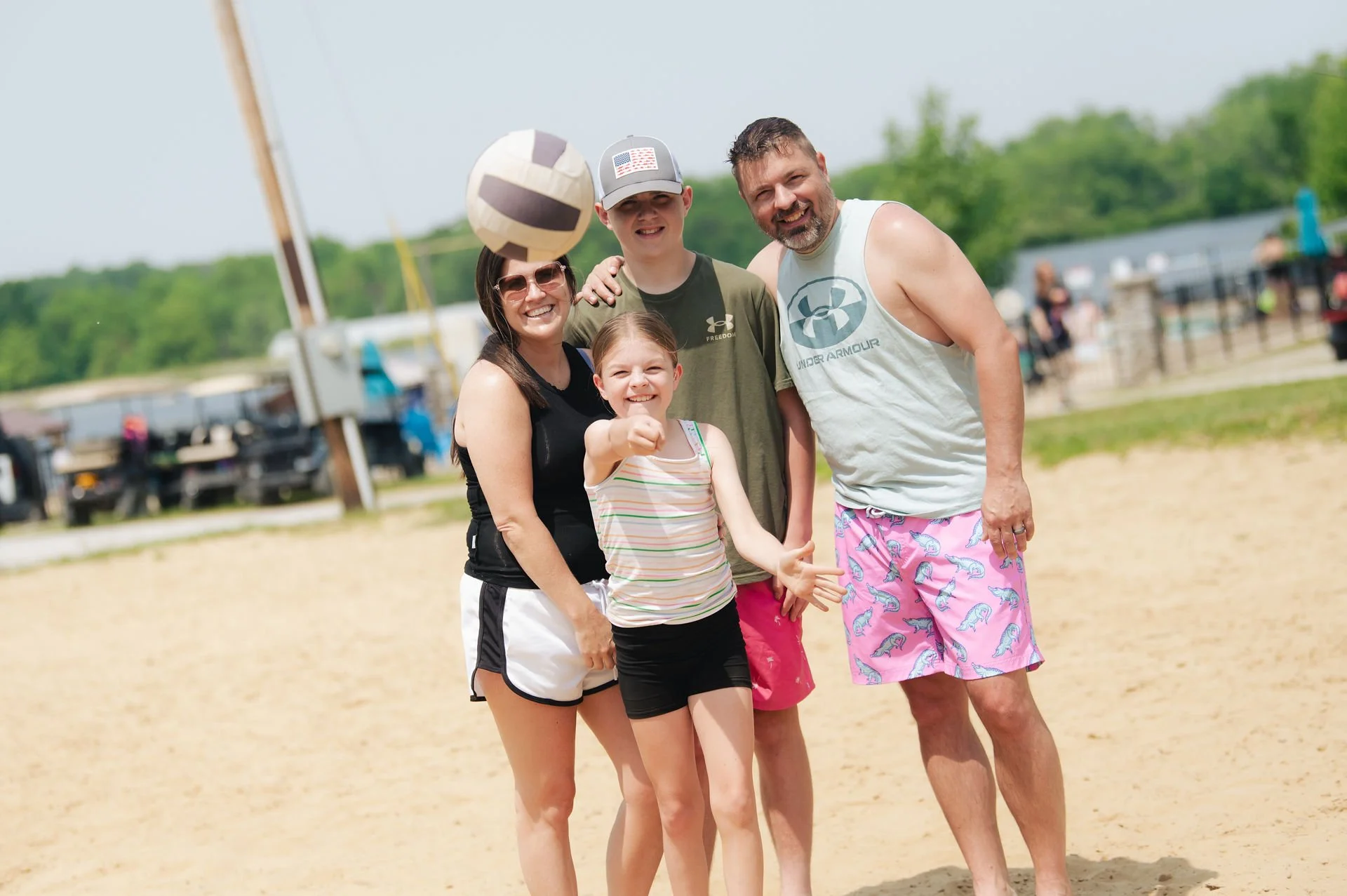 A family of five posing and smiling on a sandy beach during summer, with a volleyball bouncing above their heads.