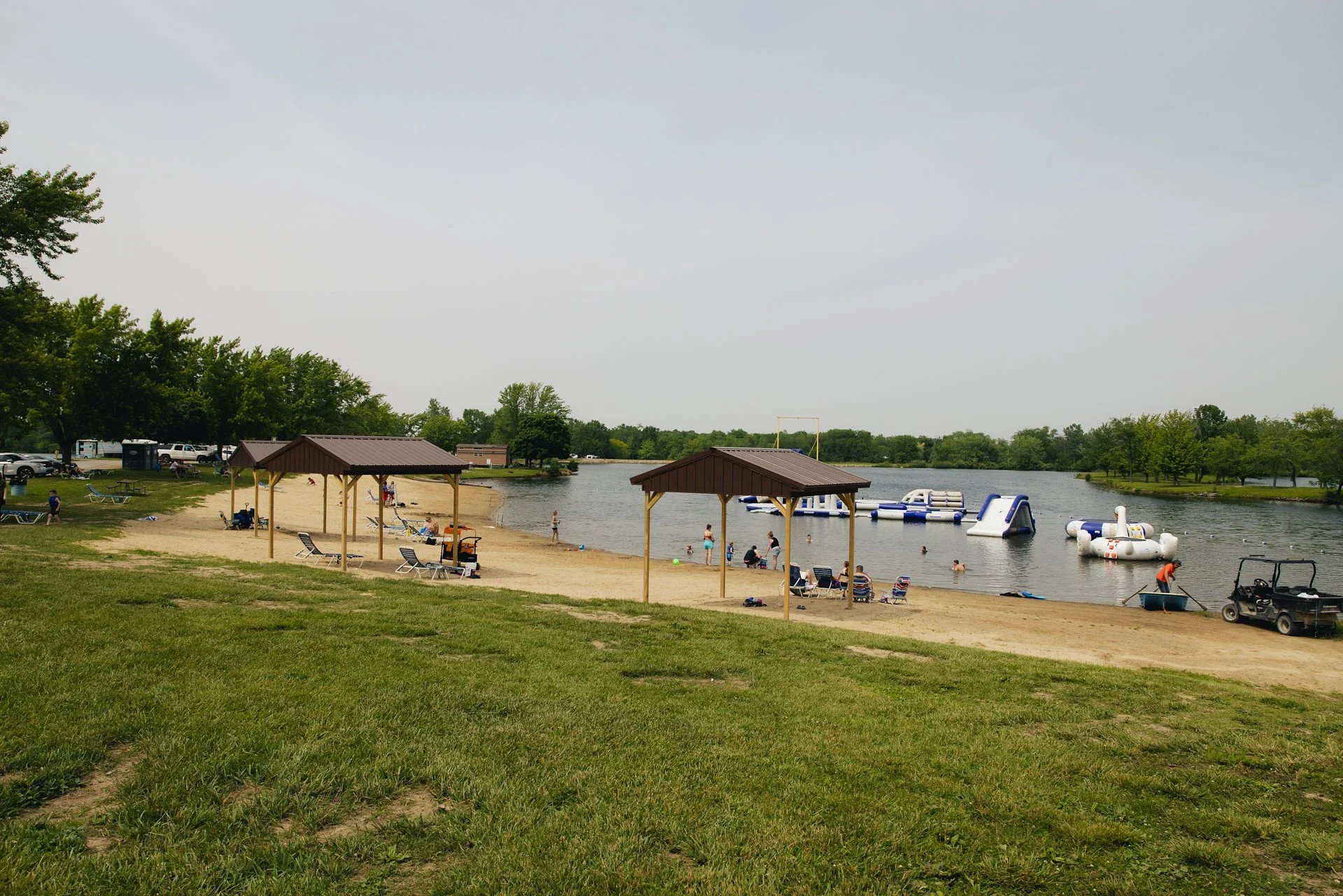 People relaxing and swimming at a lakeside beach with inflatable water structures, shaded sitting areas, and green trees in the background.
