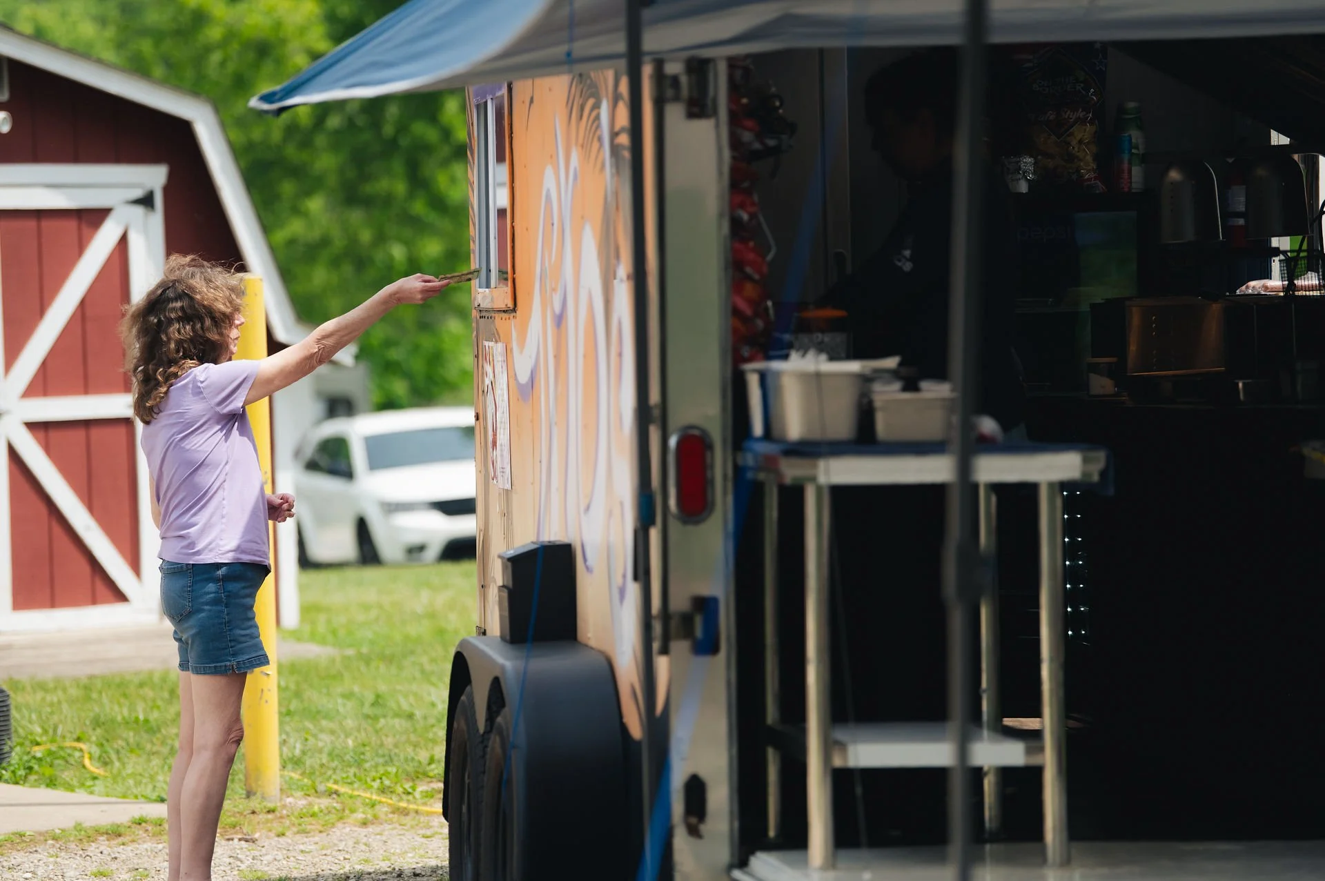 A woman with curly brown hair, wearing a light purple T-shirt and denim shorts, is paying at a food truck window with her hand reaching out holding money. The food truck has orange and white design elements and is parked outdoors on a grassy area wit