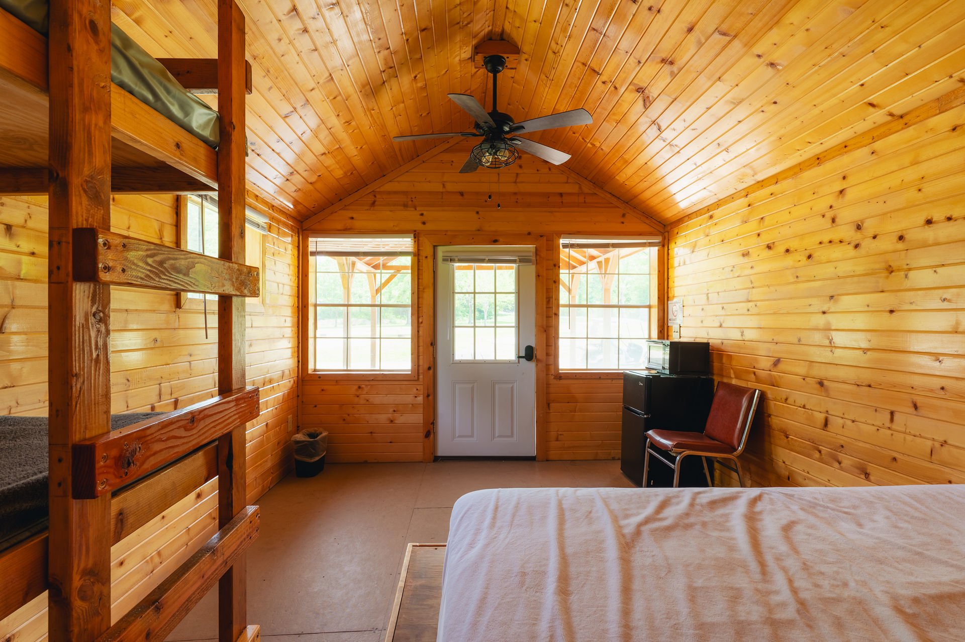 Interior of a cozy wooden cabin bedroom with windows and a ceiling fan, featuring a bunk bed, a chair, and a small refrigerator with a microwave on top.