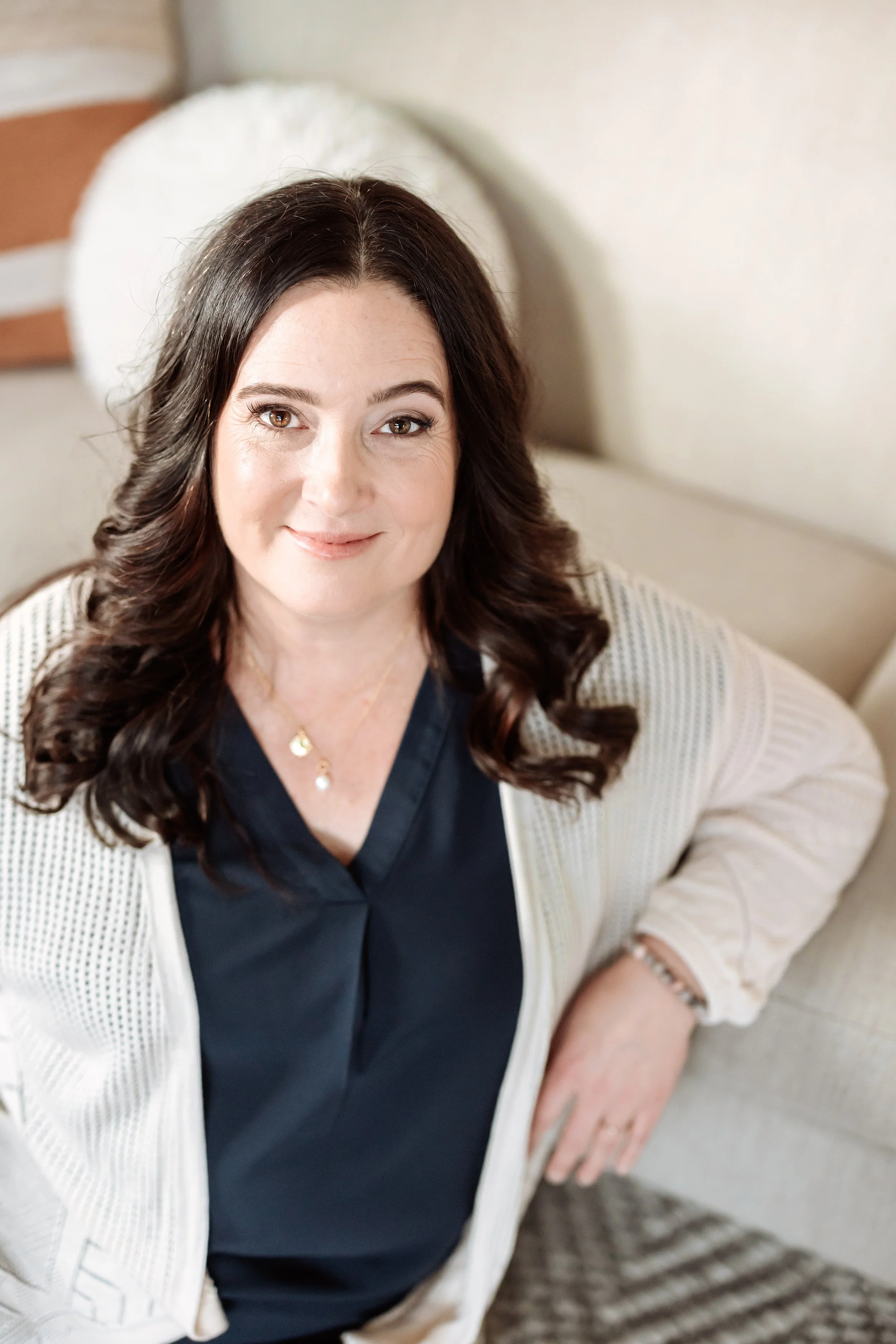 A woman with dark brown, wavy hair, wearing a navy blouse and a white cardigan, sitting on a beige couch with a white pillow behind her, smiling and looking at the camera.