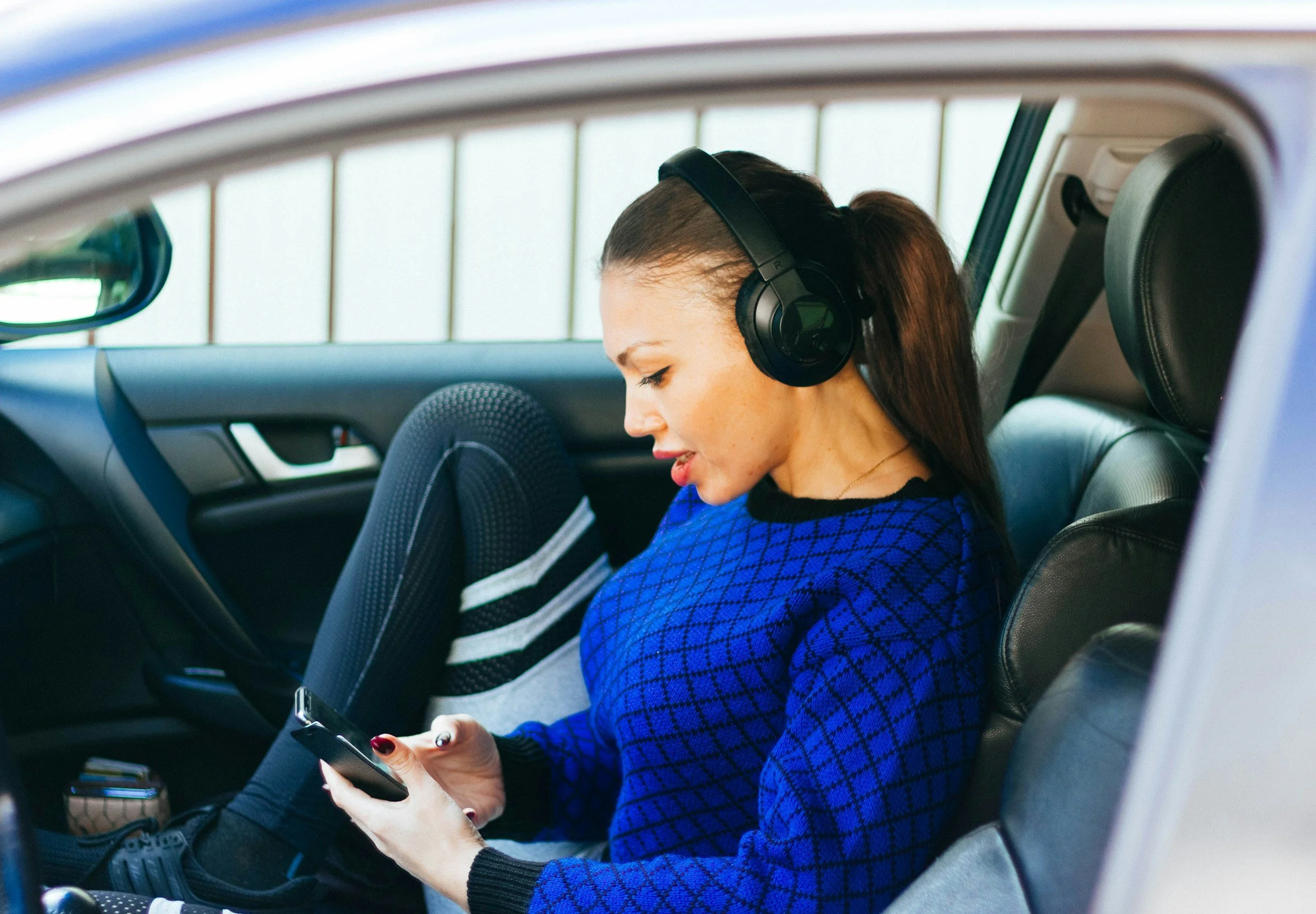 Woman in blue sweatshirt with headphones on looking at her phone while sitting in a car.